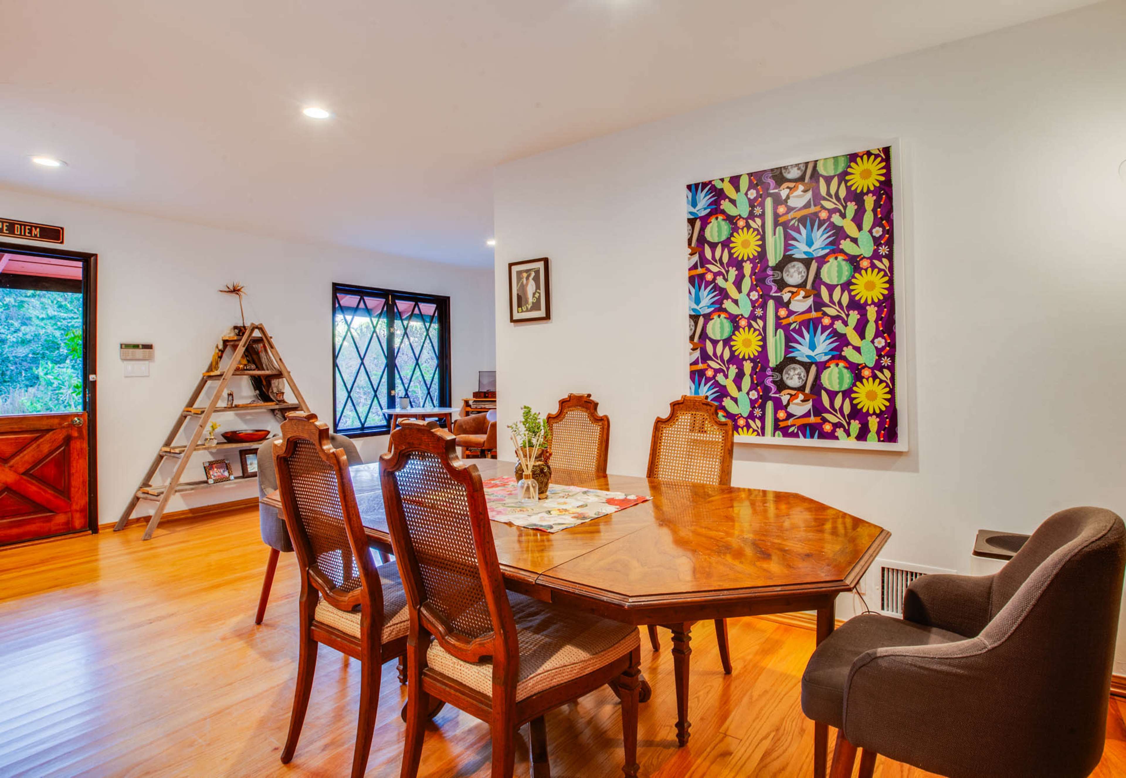 The image shows a dining room with a wooden table surrounded by eight chairs, a colorful artwork on the wall, and a decorative ladder shelf in the corner.