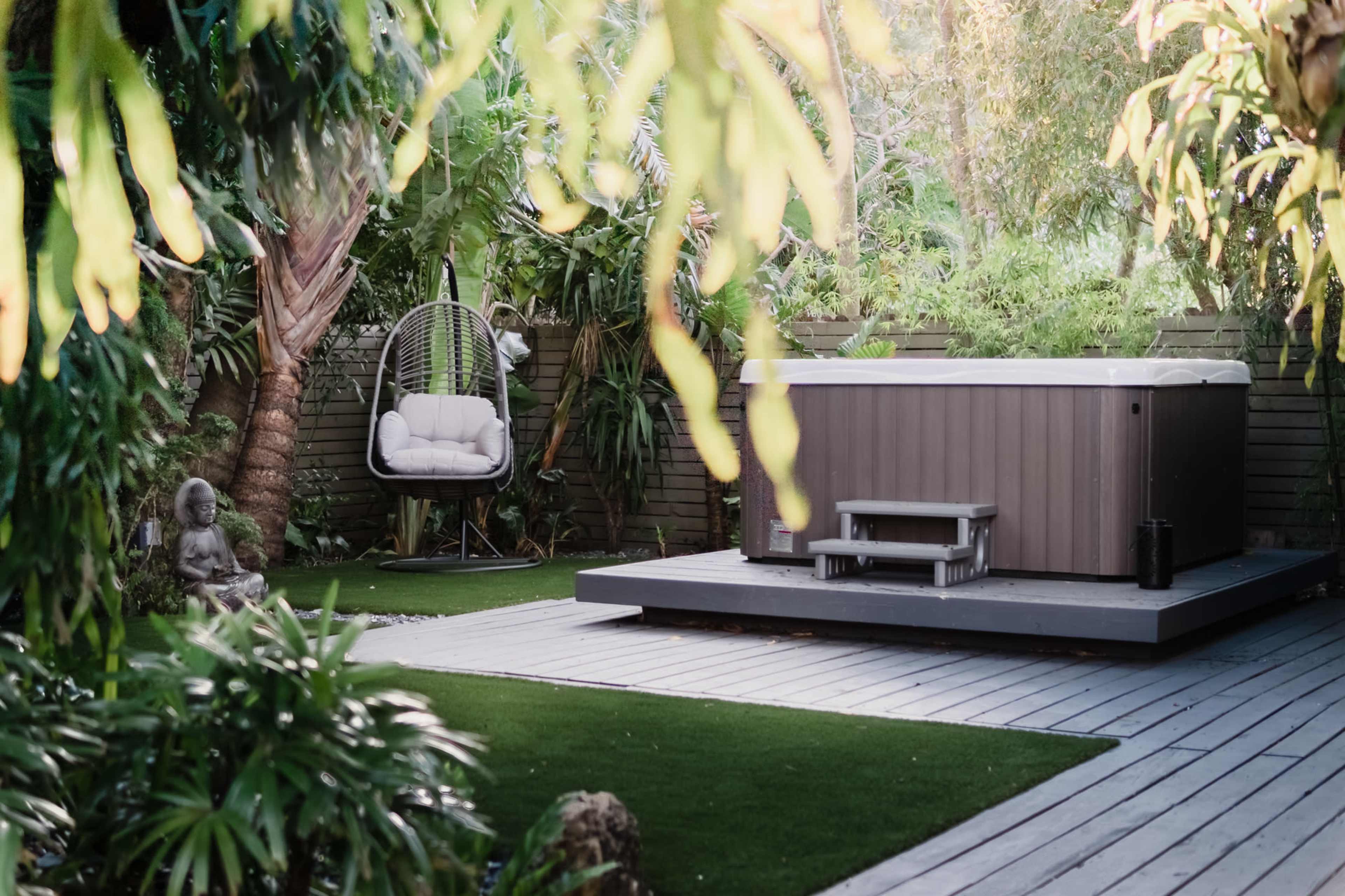A hot tub sits on a wooden deck surrounded by lush greenery and a hanging chair in a tranquil garden setting.