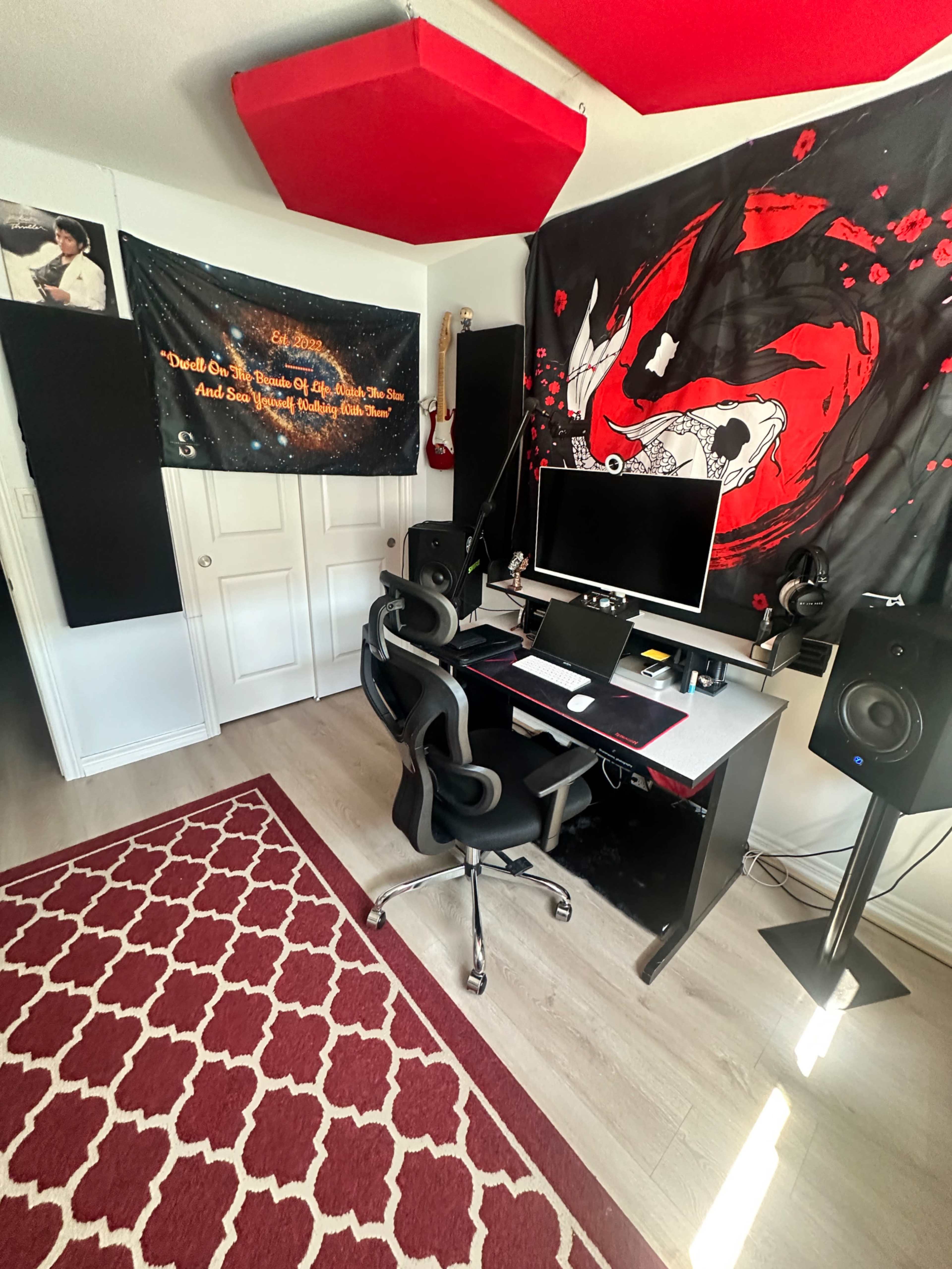 The image shows a home studio setup with a desk, computer monitor, speakers, and acoustic panels on the walls, alongside decorative banners and a patterned rug.