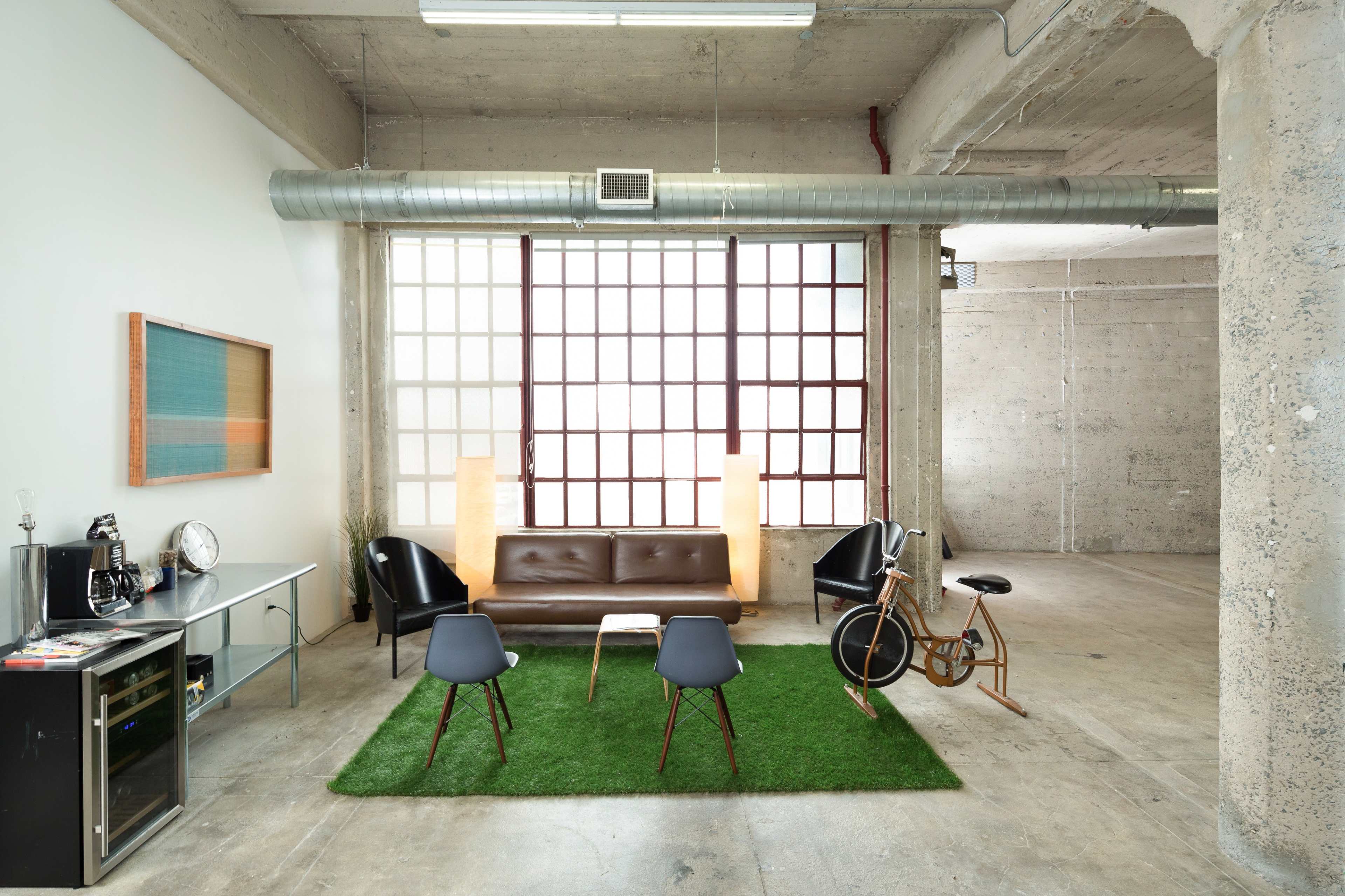 A minimalist living space with a brown leather sofa, two gray chairs, a coffee table on green grass-like carpet, and large windows that let in natural light.