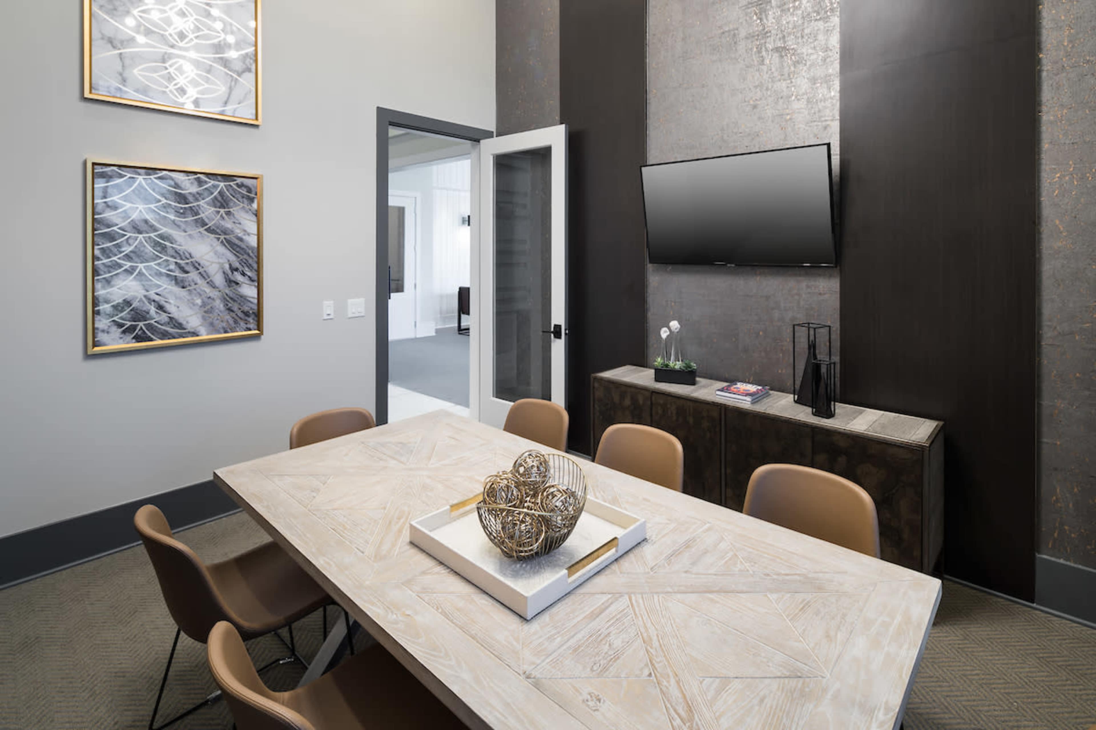 A modern conference room features a wooden table surrounded by brown chairs, with abstract art on the walls and a television mounted on a dark accent wall.
