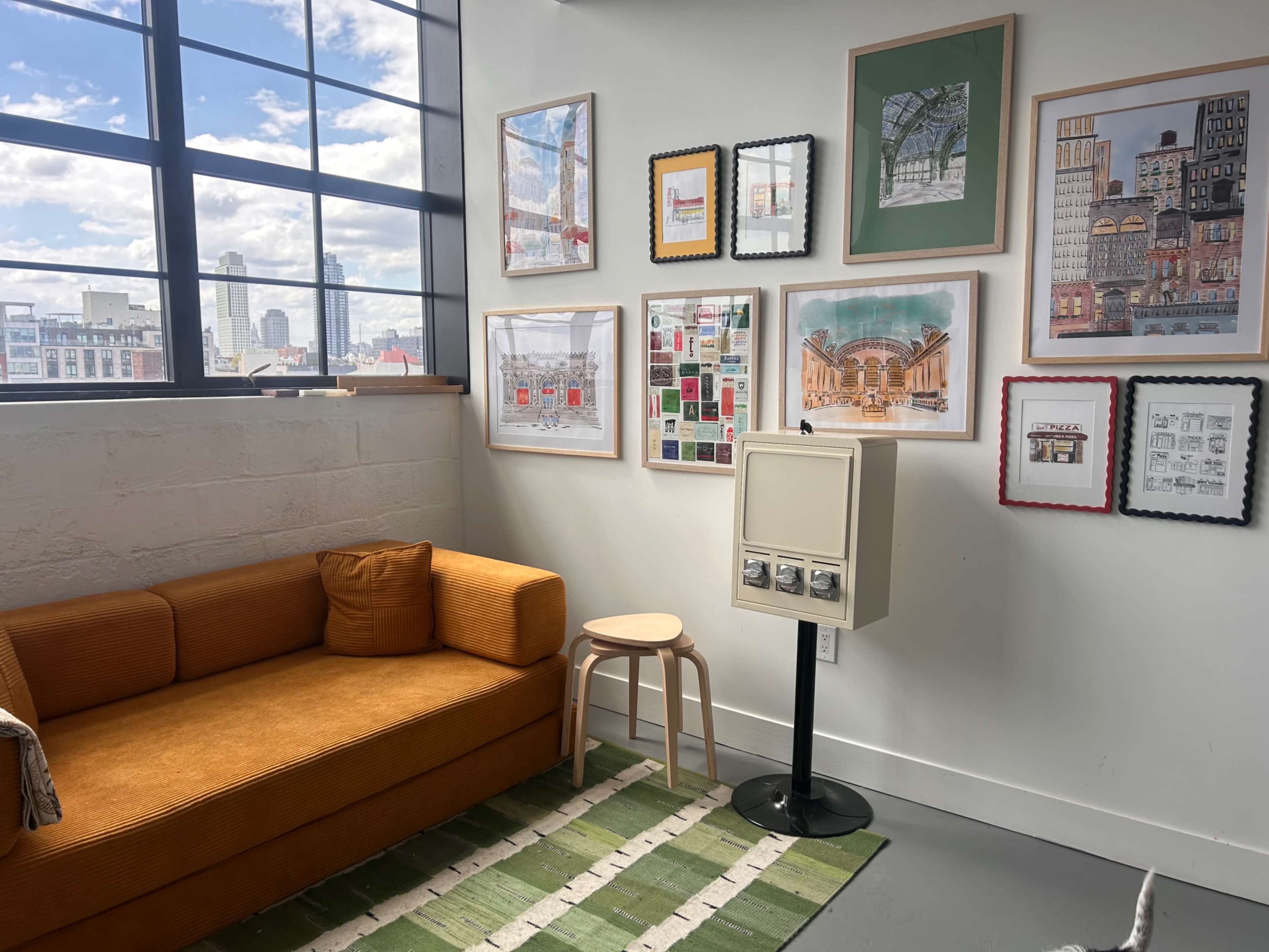 The image shows a cozy corner with an orange couch, a stool, a green striped rug, and various framed artworks on the wall beside a large window.