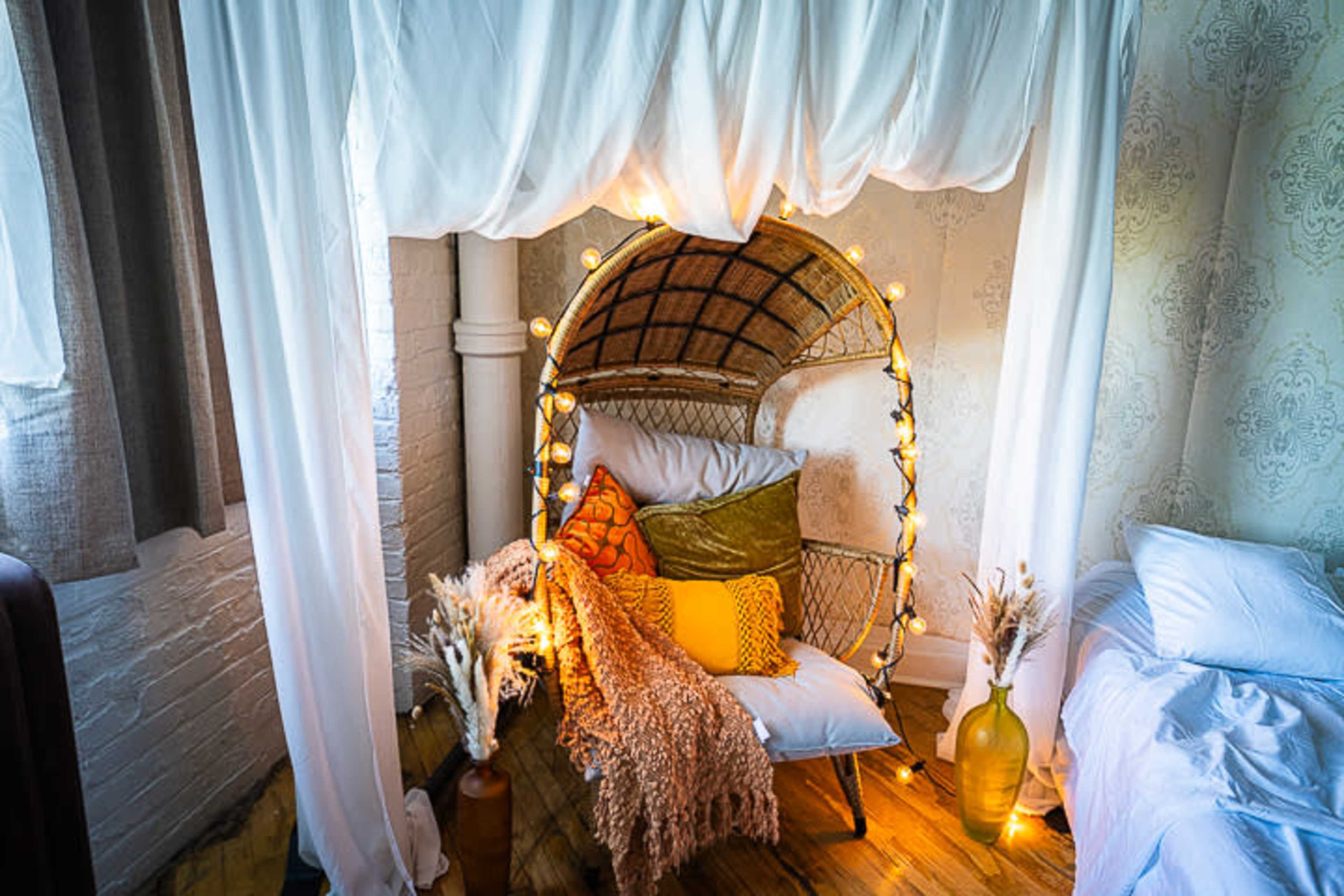 A cozy nook features a rattan chair draped in fairy lights and surrounded by decorative pillows and a soft throw blanket.
