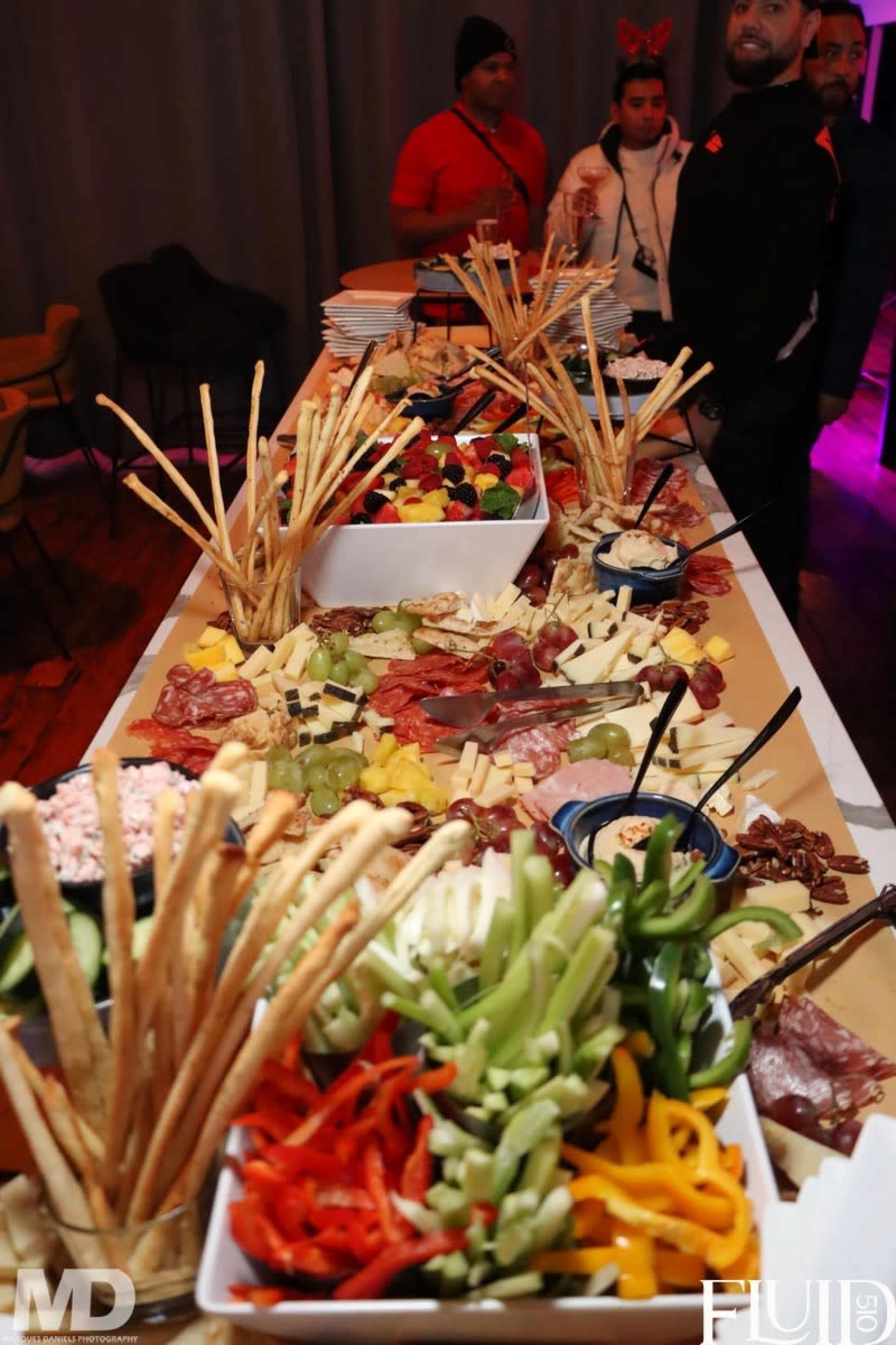 A long buffet table is adorned with a variety of cheeses, meats, vegetables, and breadsticks arranged in colorful displays.