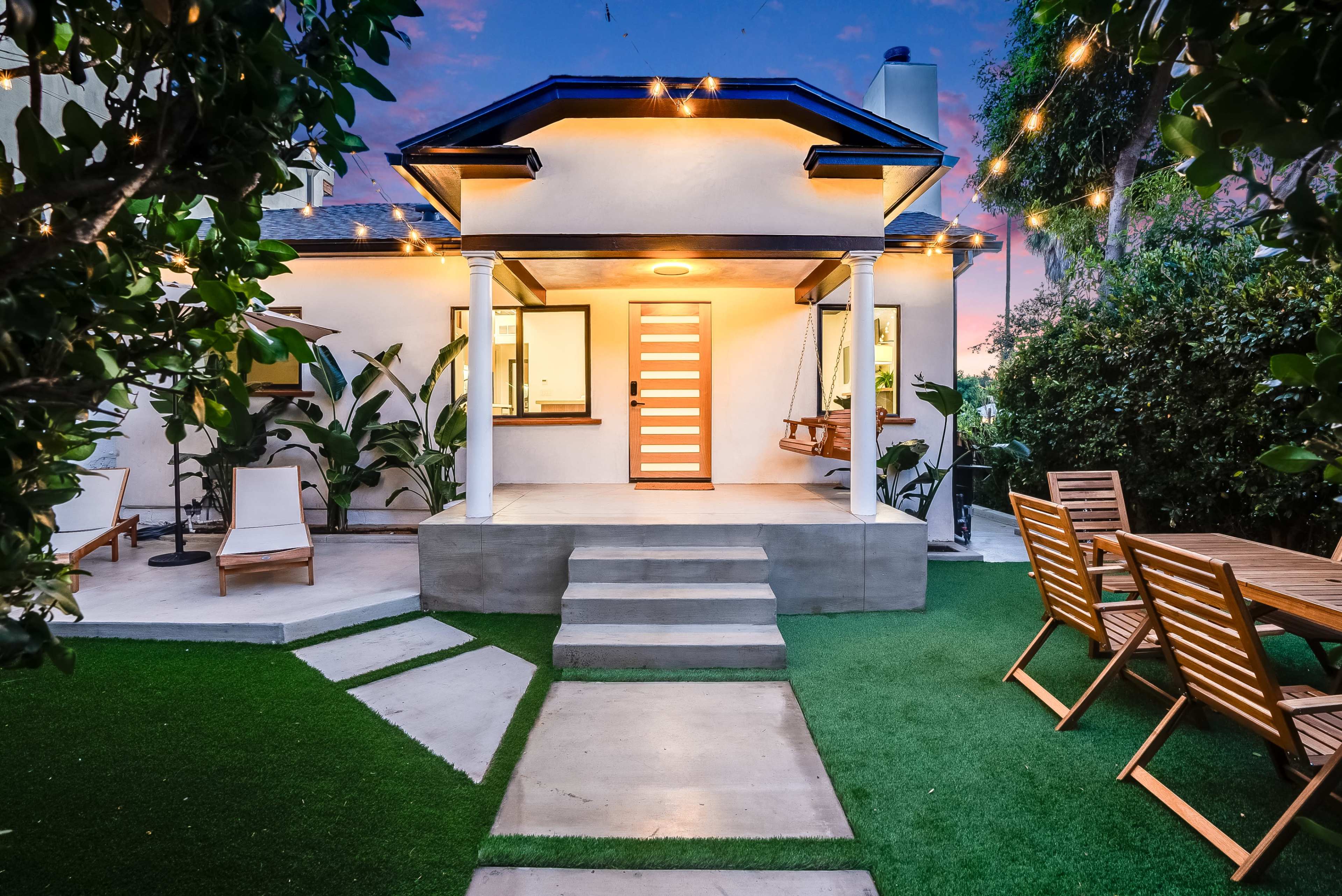 The image shows a modern house with a front porch, white exterior, and a wooden door, surrounded by landscaped greenery and outdoor furniture.