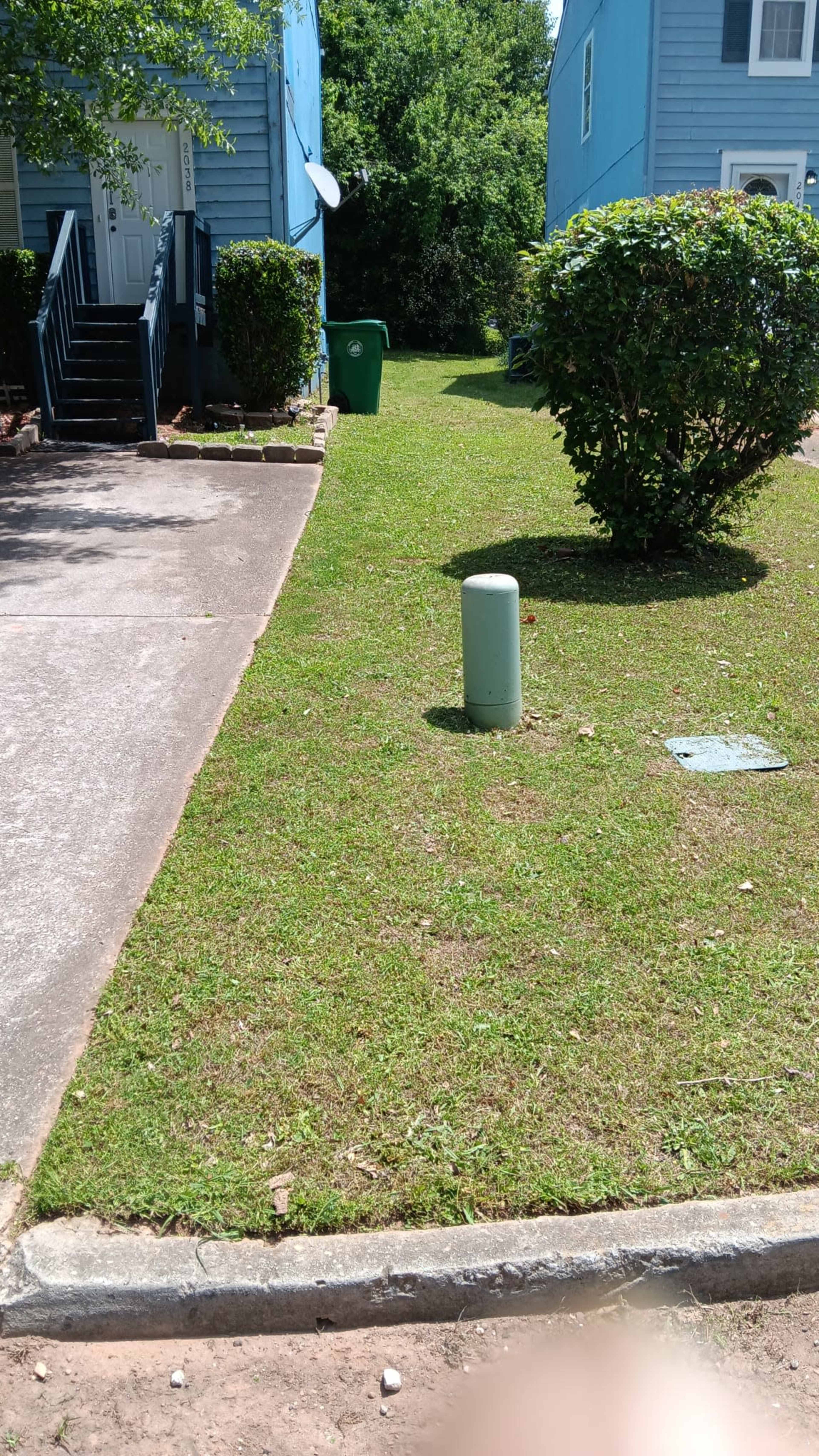 A grassy area between two blue houses, with a green trash bin and a concrete pole near the edge of the driveway.
