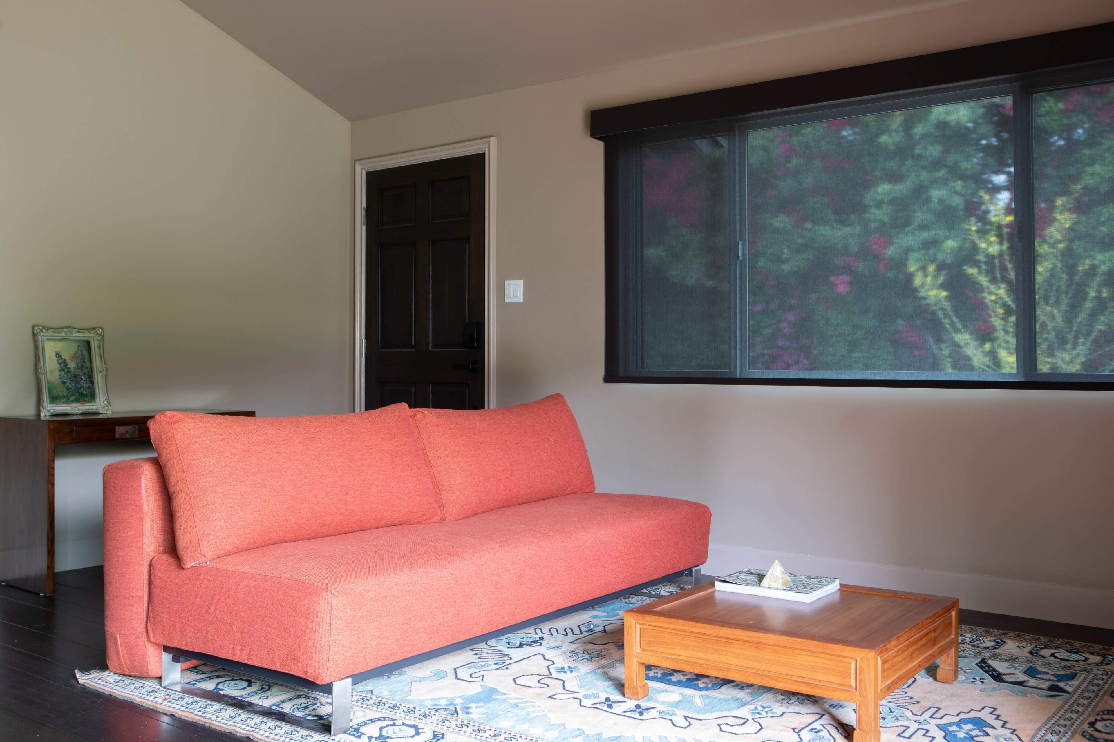 The image shows a modern living room with an orange sofa, a wooden coffee table, and a large window overlooking greenery.