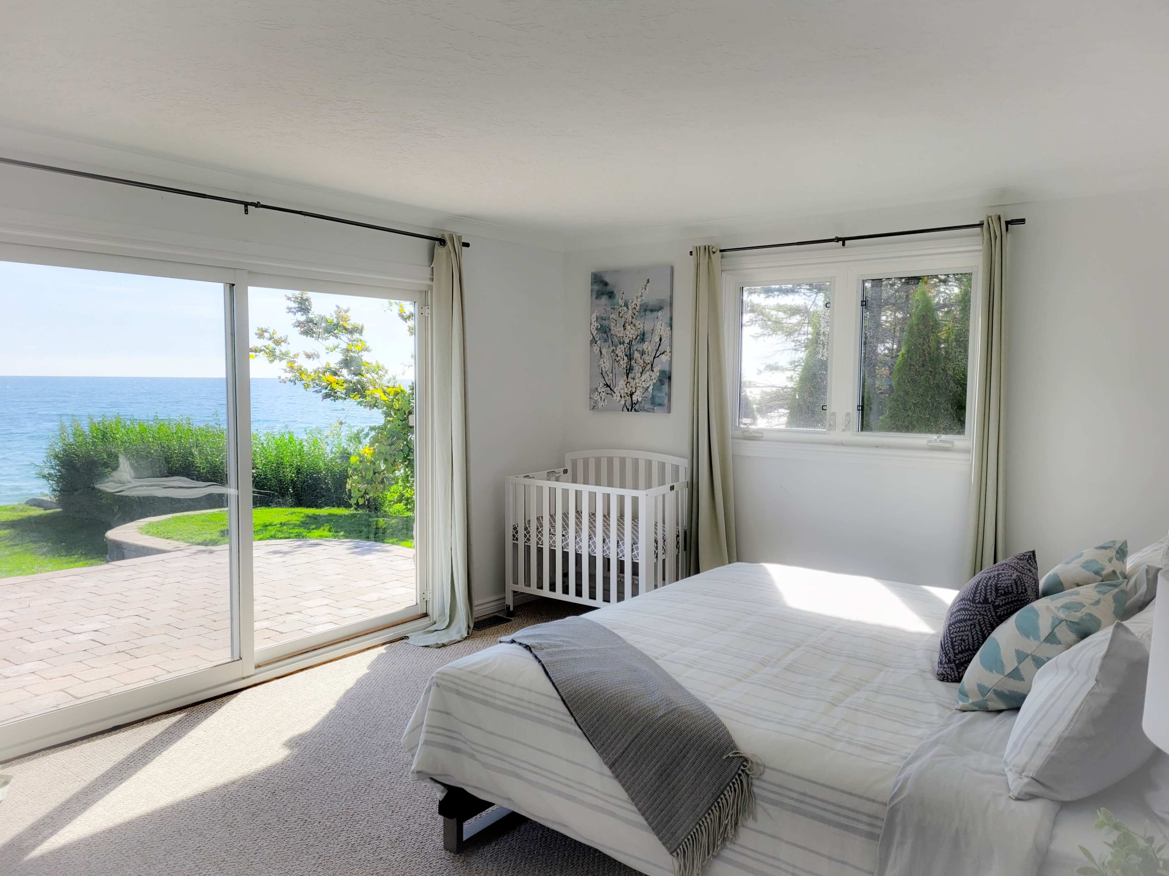 A bright bedroom features a crib next to a large window overlooking a view of the ocean and lush greenery outside.
