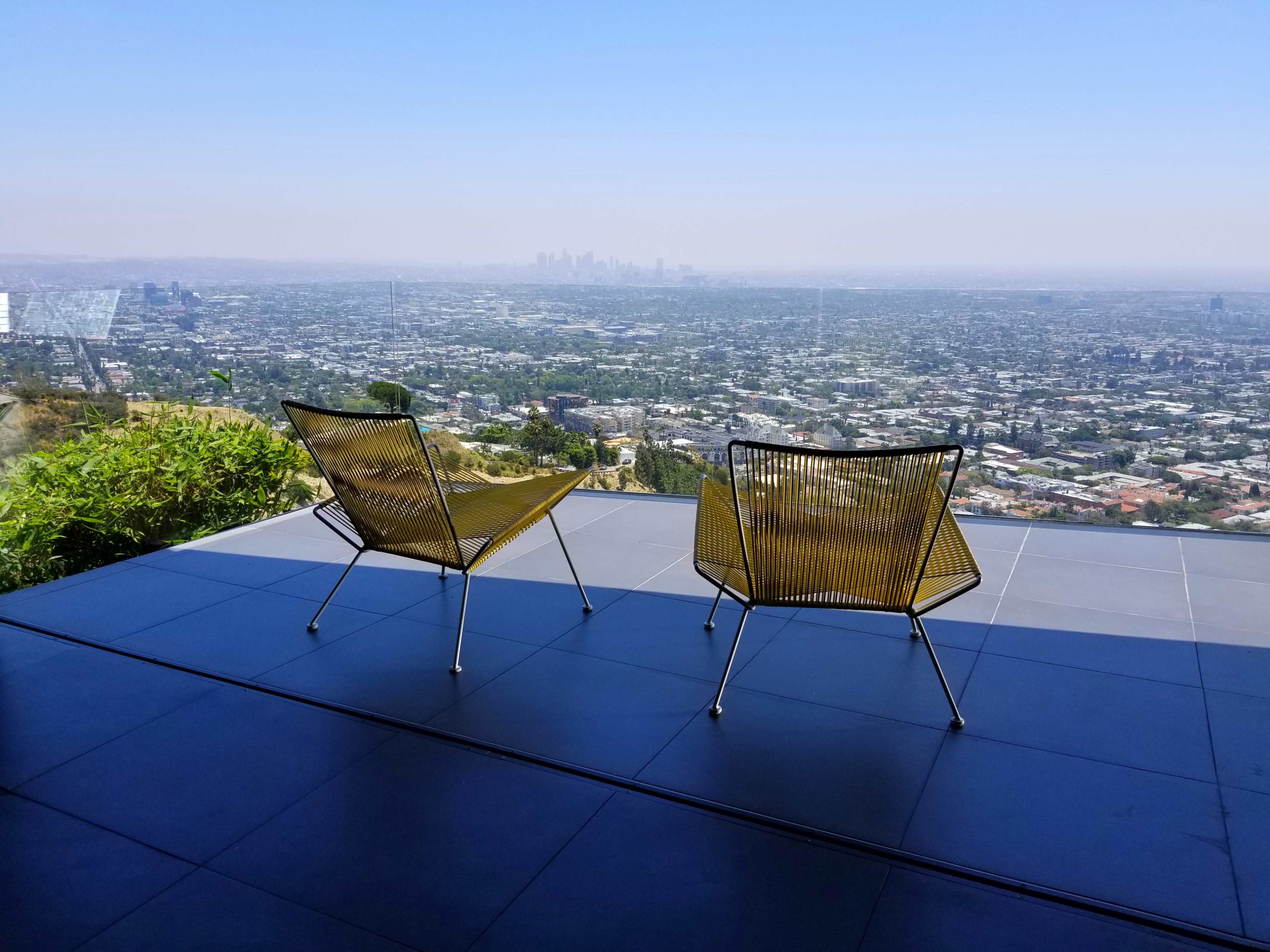 Two yellow wire chairs are positioned on a patio overlooking a sprawling view of a cityscape.
