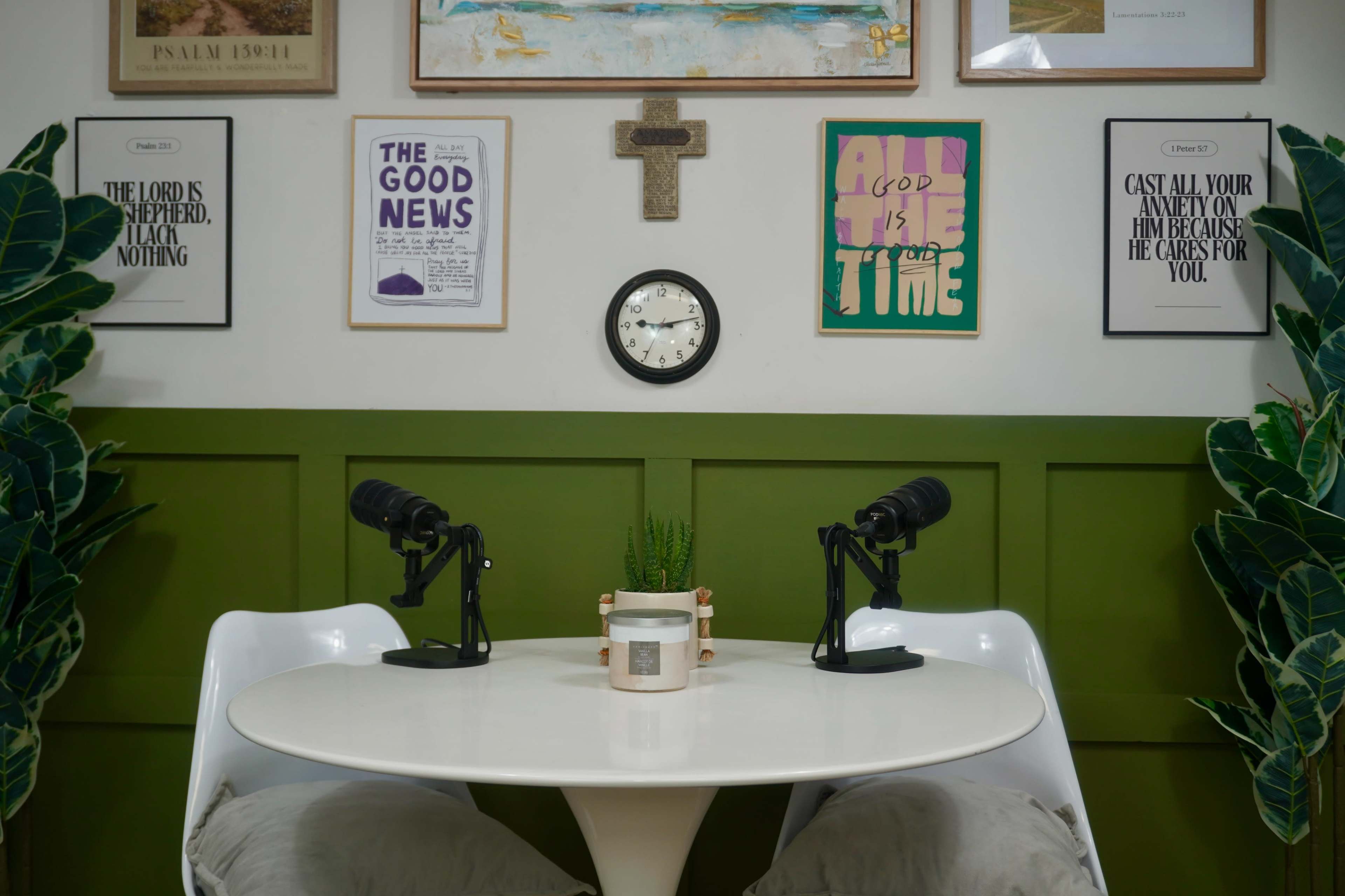 The image shows a small, round white table with two chairs, adorned with microphones, surrounded by framed artwork and decorative plants against a green wall.