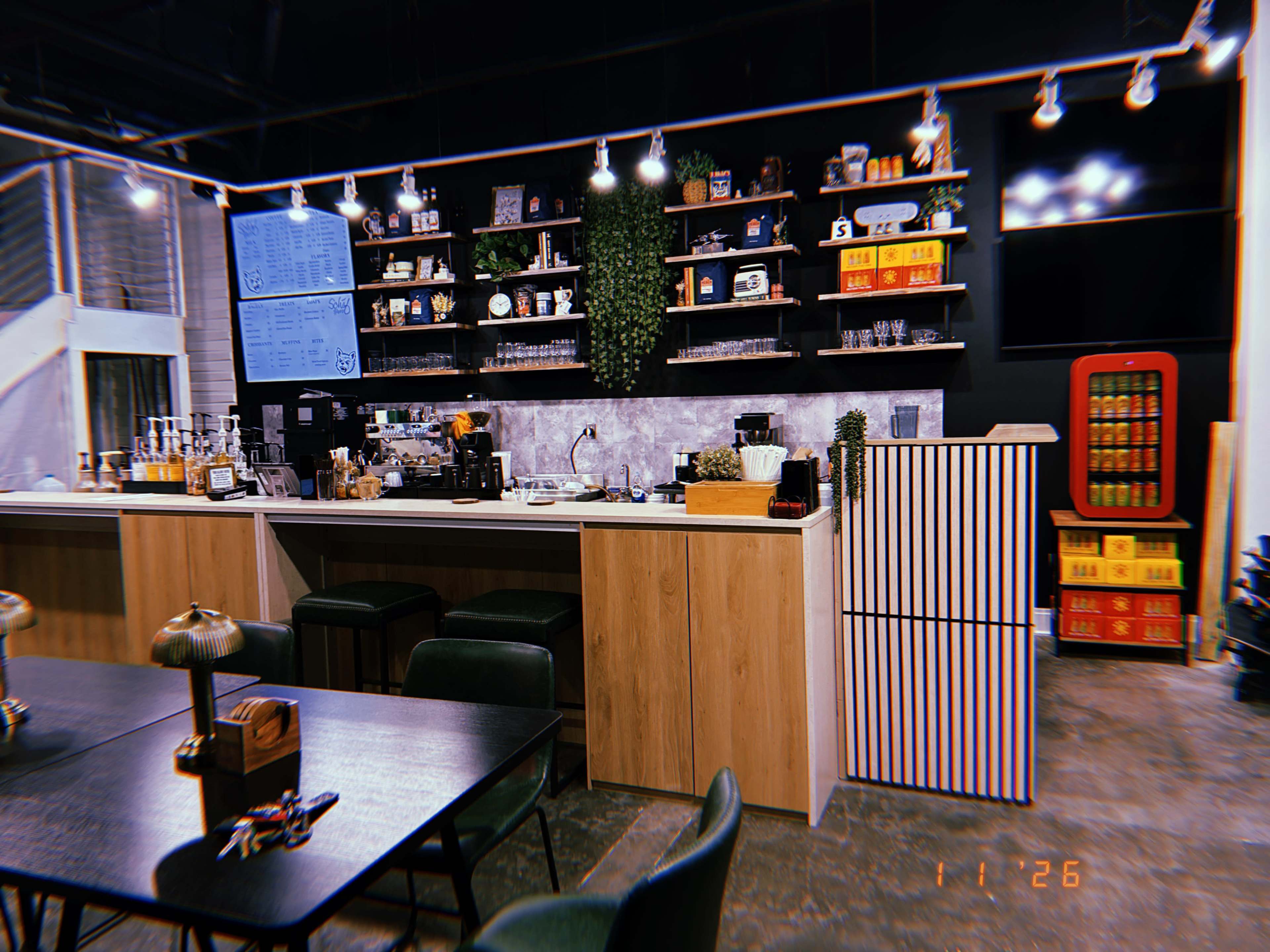The image displays a modern coffee shop interior featuring a wooden counter, shelves filled with various coffee-related items, and a seating area with dark tables and green chairs.
