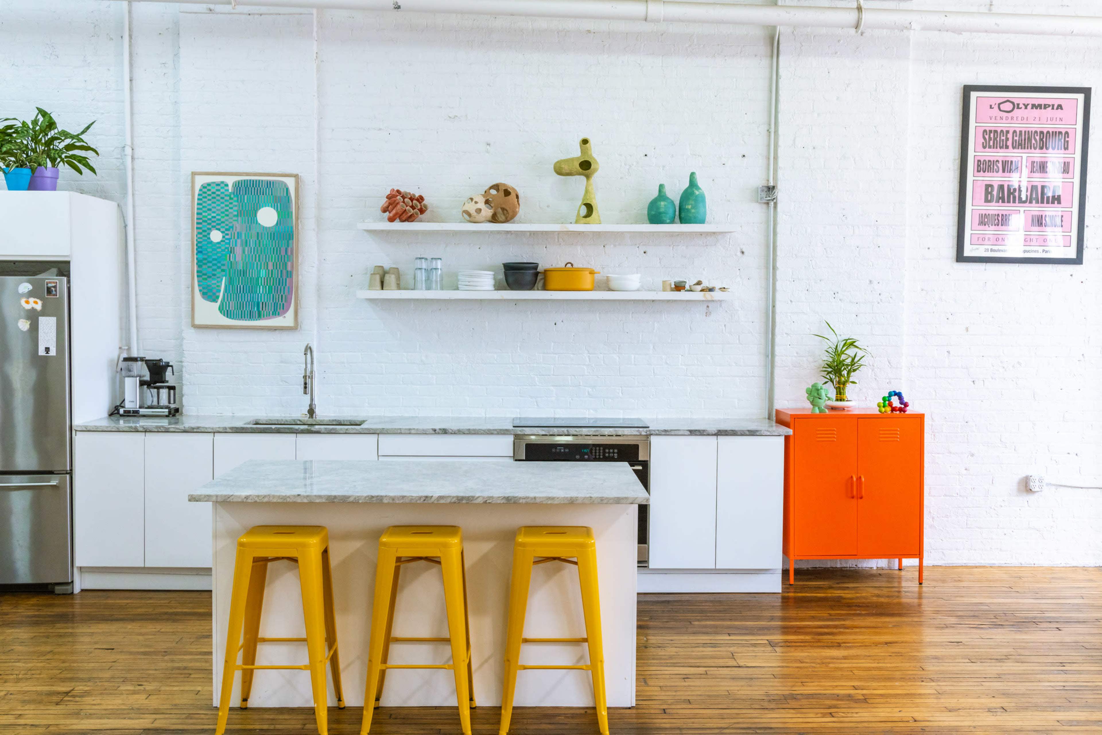 A modern kitchen features a white brick wall, an island with yellow stools, and various decorative items on shelves.