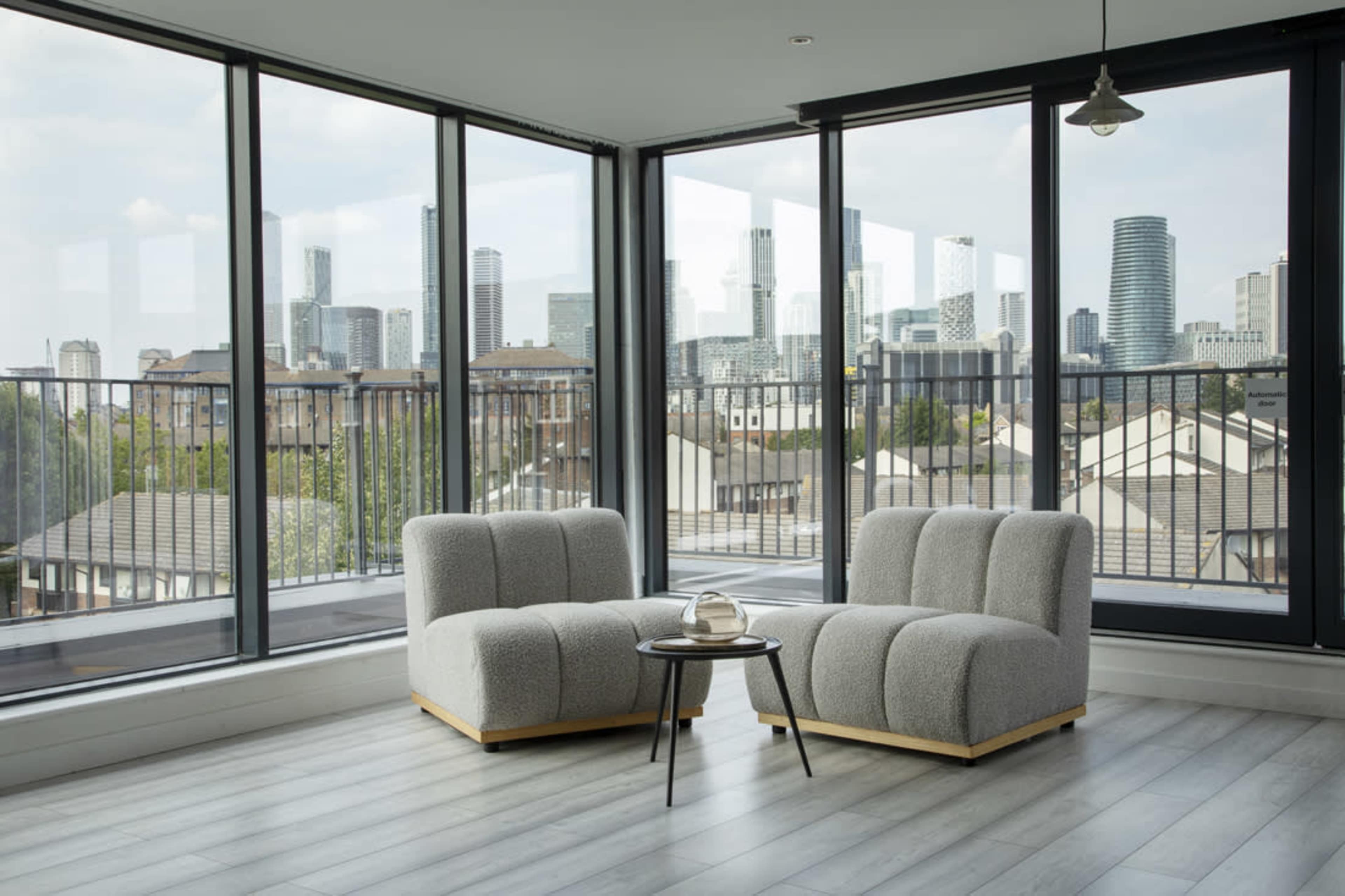 Two gray upholstered chairs with a small black side table are positioned near large windows offering a view of a city skyline.