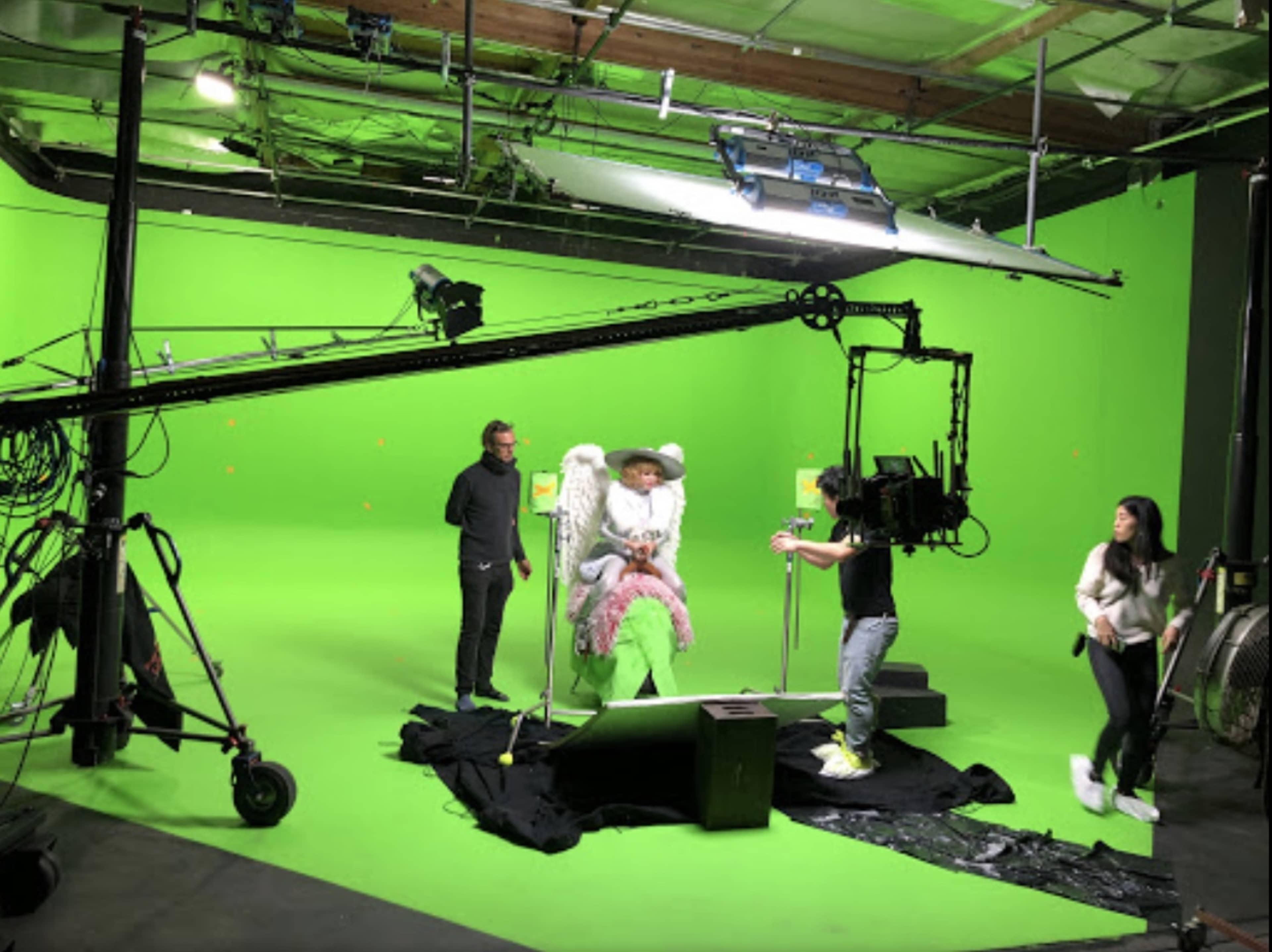 A production crew sets up a green screen studio with a cameraman filming a person dressed in costume seated on a large chair.