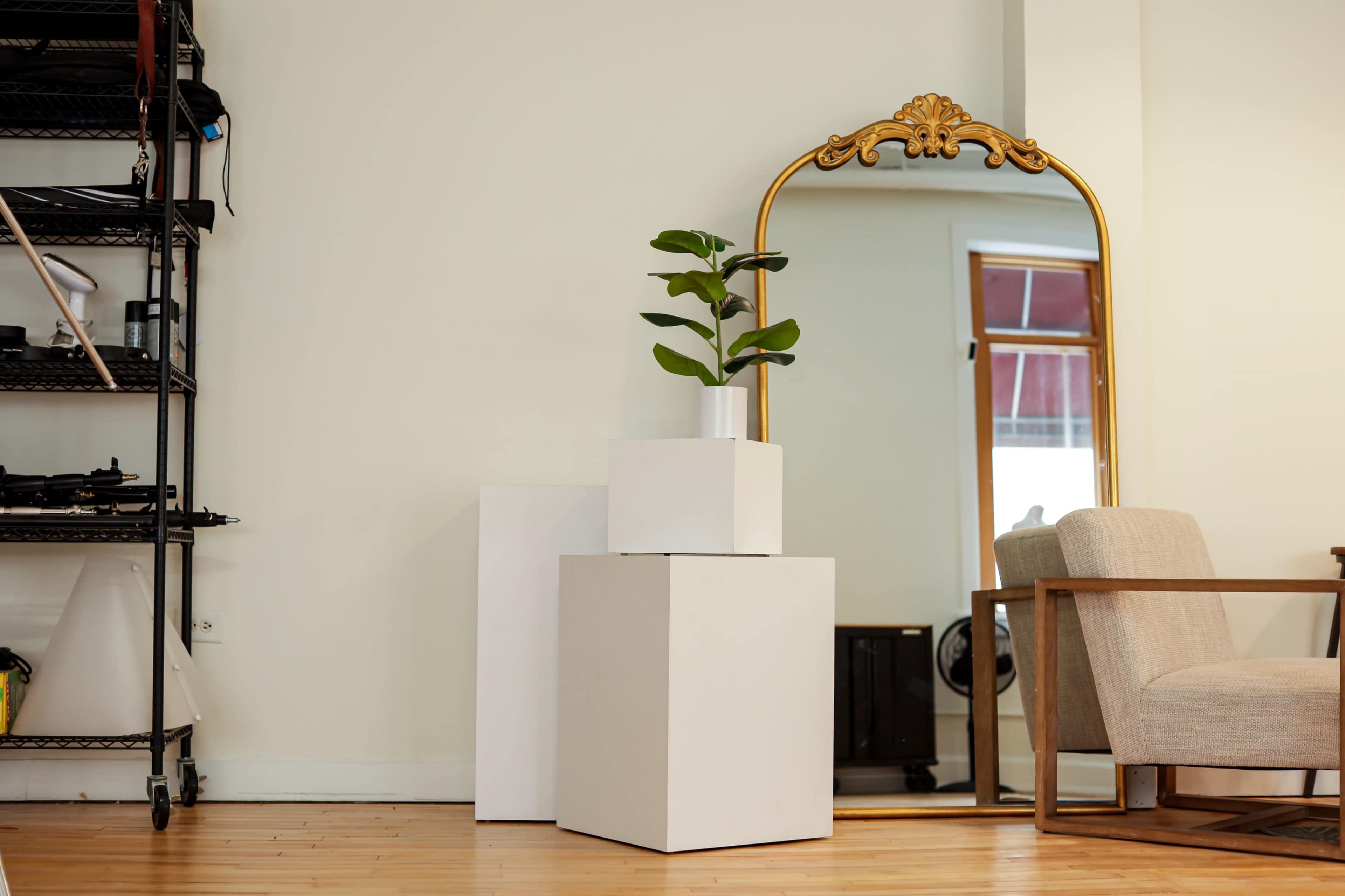 The scene features a minimalist interior with stacked white cubes, a potted plant on top, a large ornate mirror, and a wooden chair.
