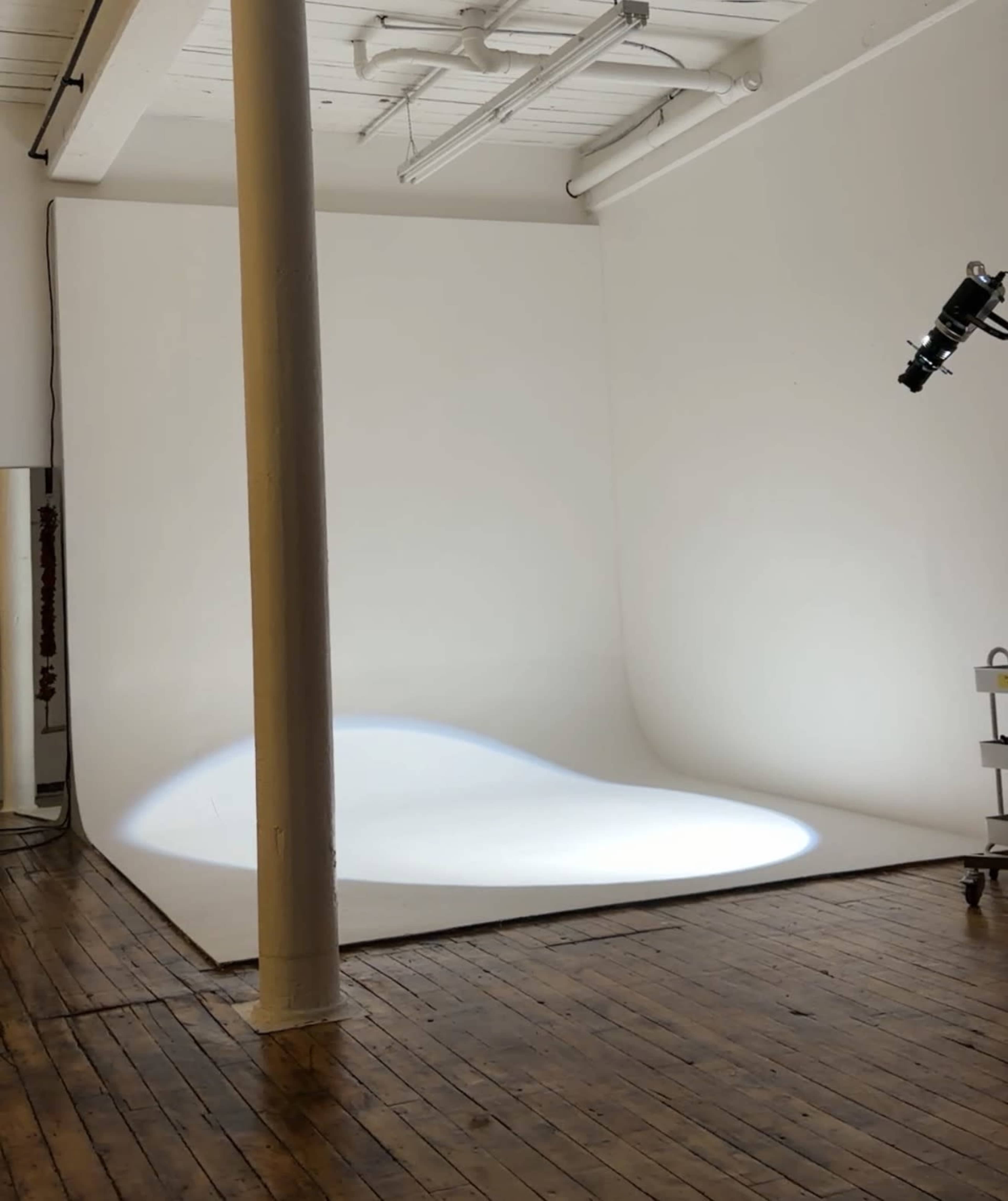 The image shows a minimalistic photo studio with a curved white backdrop, a wooden floor, and lighting equipment set up in the corner.