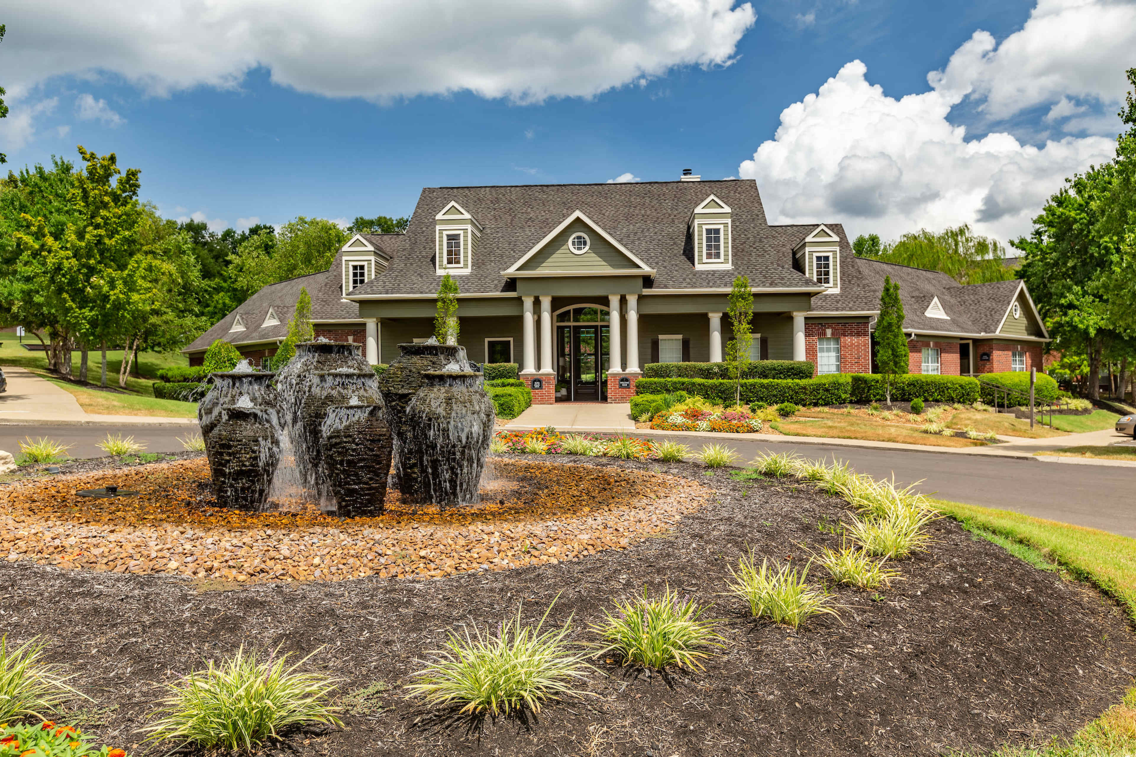 A large, modern building with a sloped roof is surrounded by a landscaped area featuring a fountain made from stacked, dark stone jars.