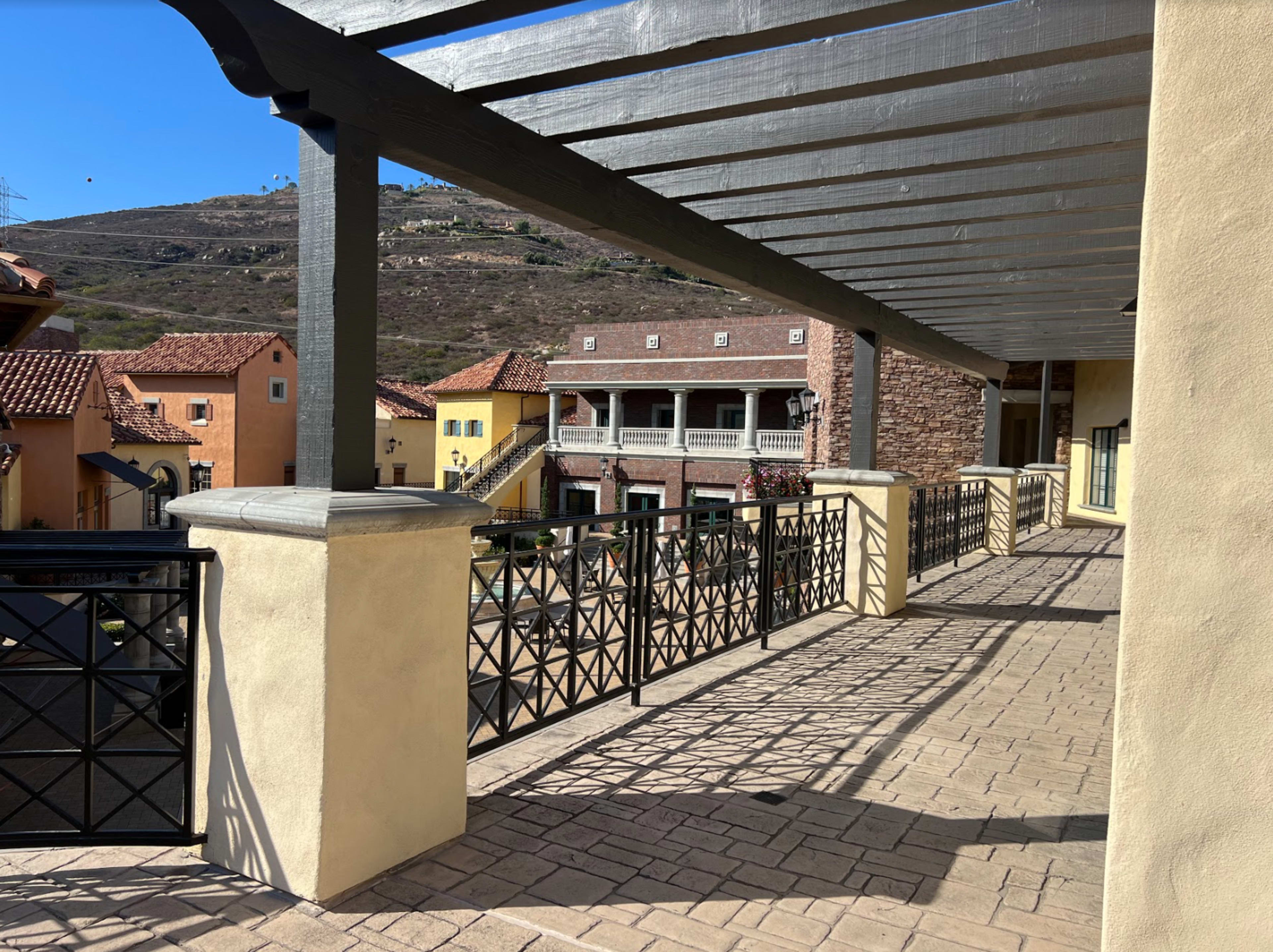 A pathway with a pergola casts shadows over a cobblestone walkway lined with buildings in a rustic architectural style.