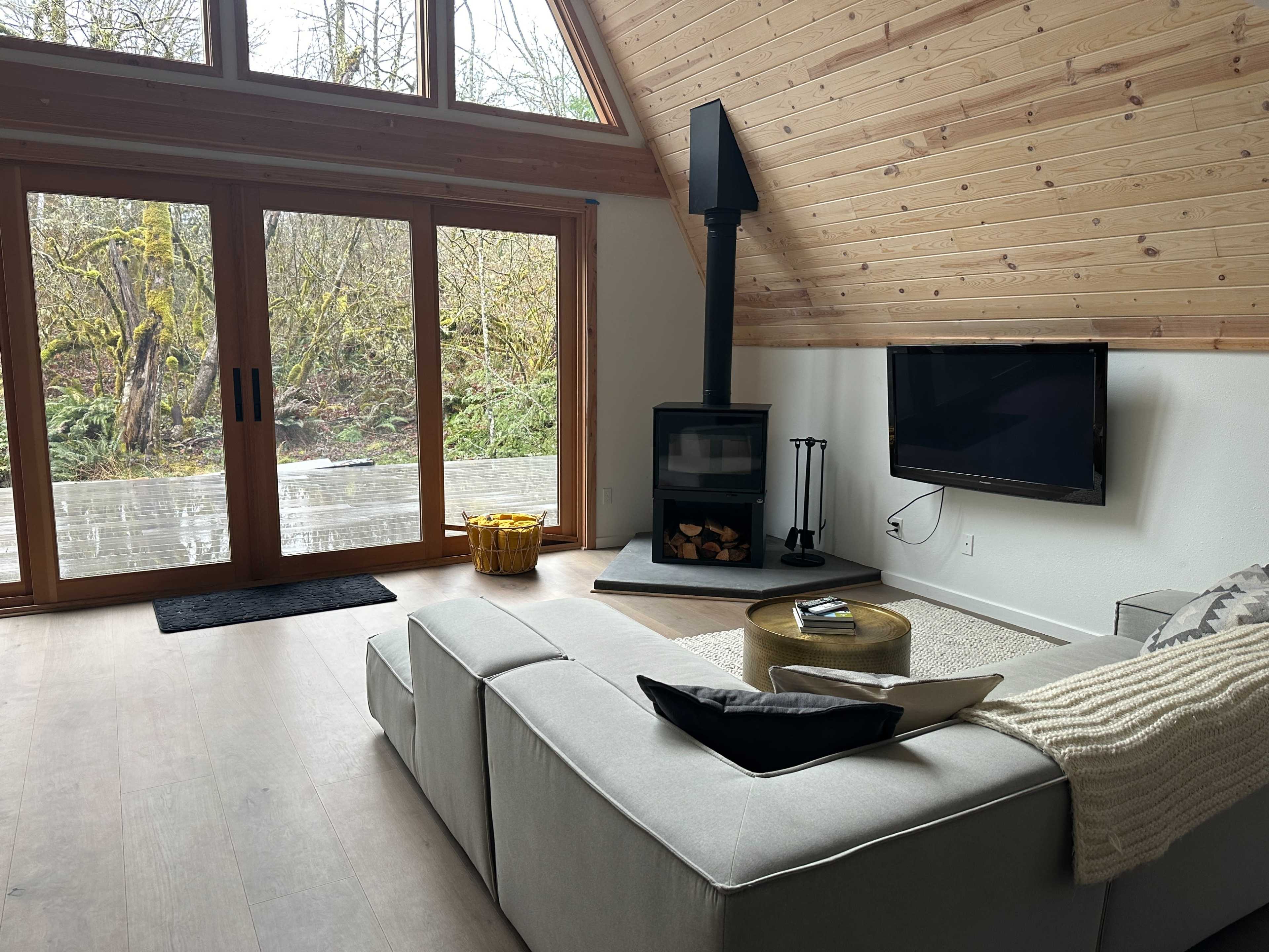 A cozy living room with a sectional sofa, a modern wood stove, a television mounted on the wall, and large sliding glass doors leading to a deck.
