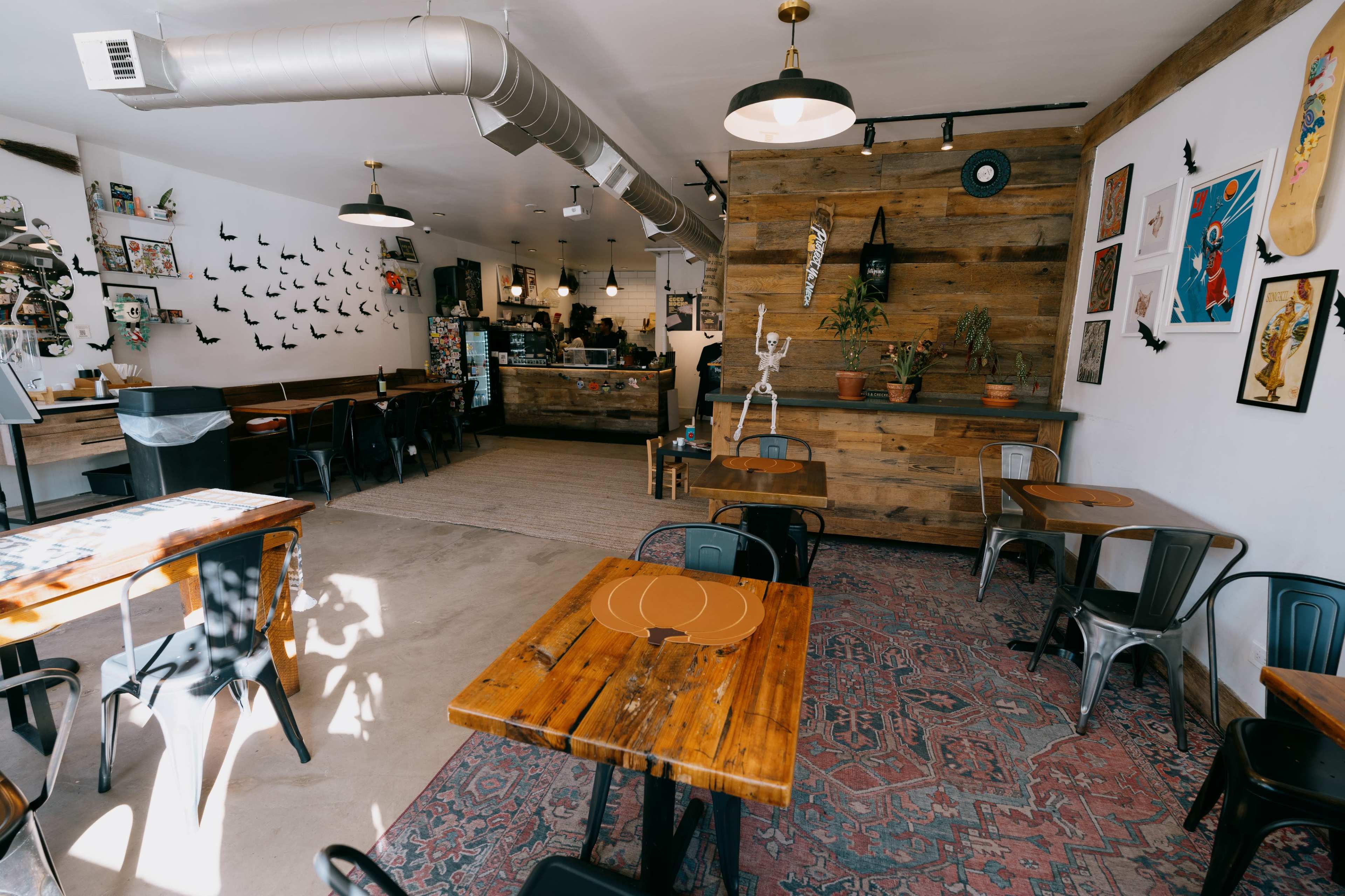The image shows a cozy café interior featuring wooden tables, metal chairs, and a mix of wall decorations, including artwork and plants.