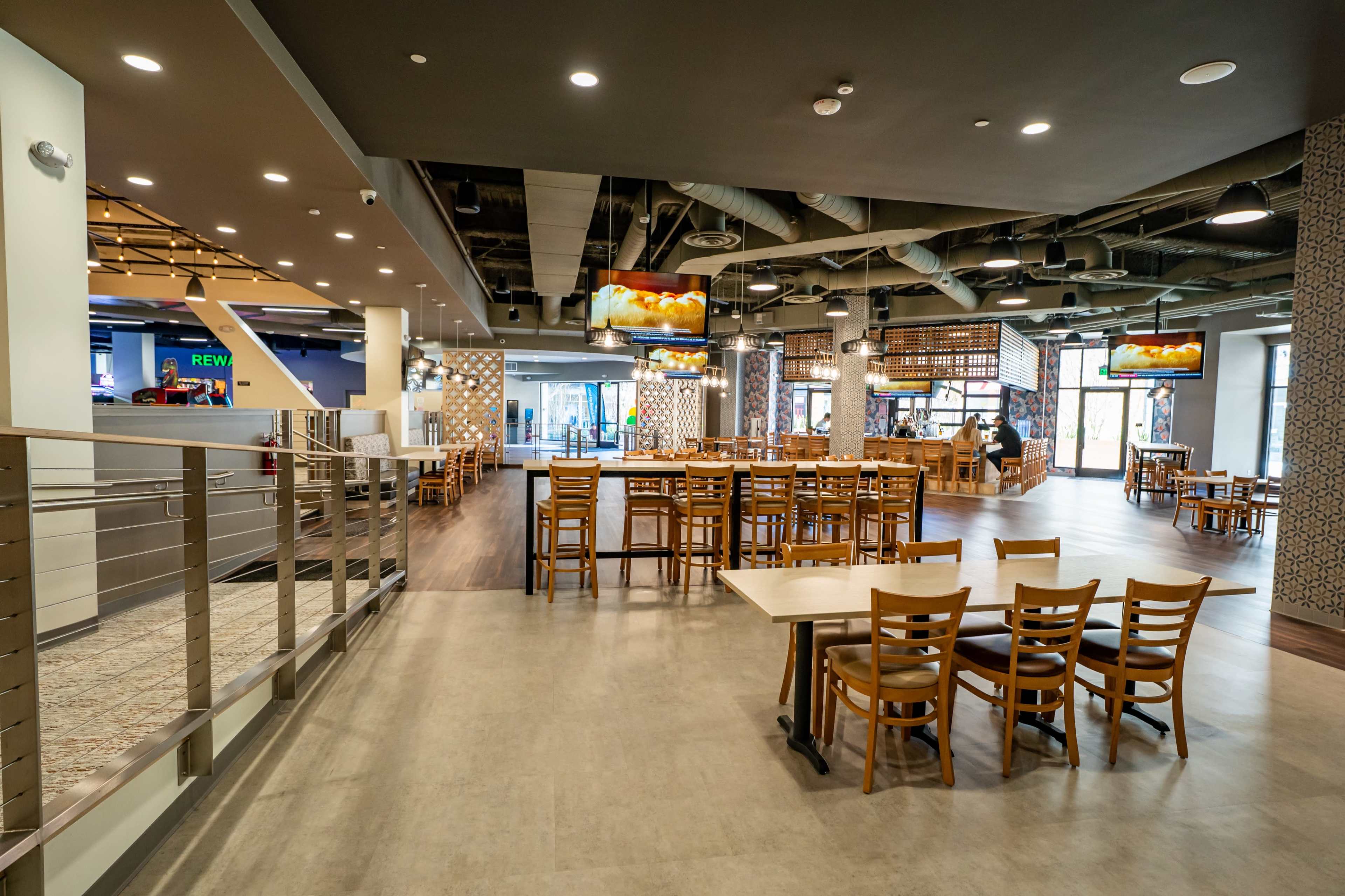 The image shows an interior view of a spacious restaurant with wooden tables and chairs, large windows, and a modern design.