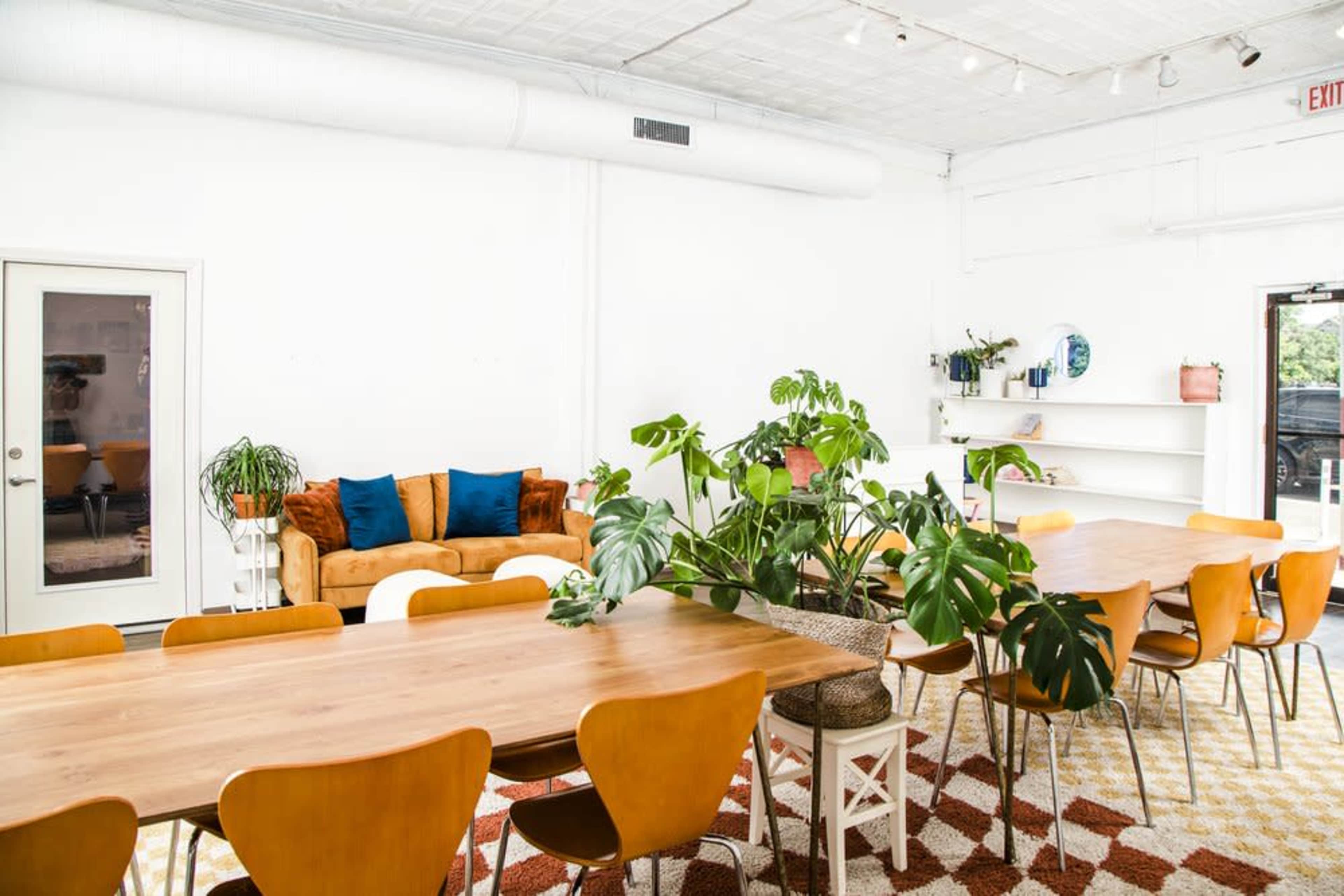 A bright, open interior space featuring a large wooden dining table, orange chairs, a sofa with blue cushions, and various plants throughout.