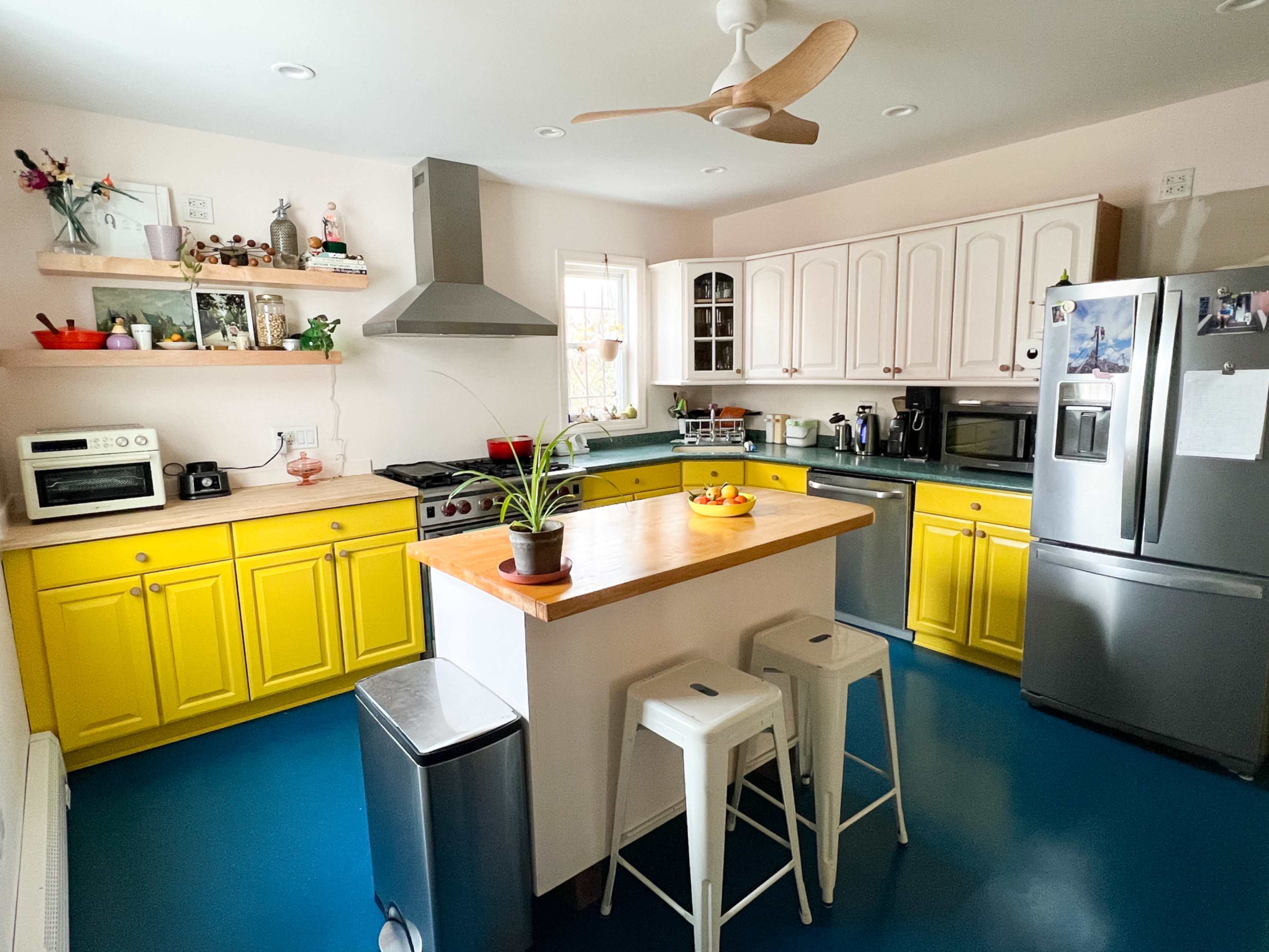 The kitchen features yellow cabinets, stainless steel appliances, a central island with a wooden countertop, and a blue floor.