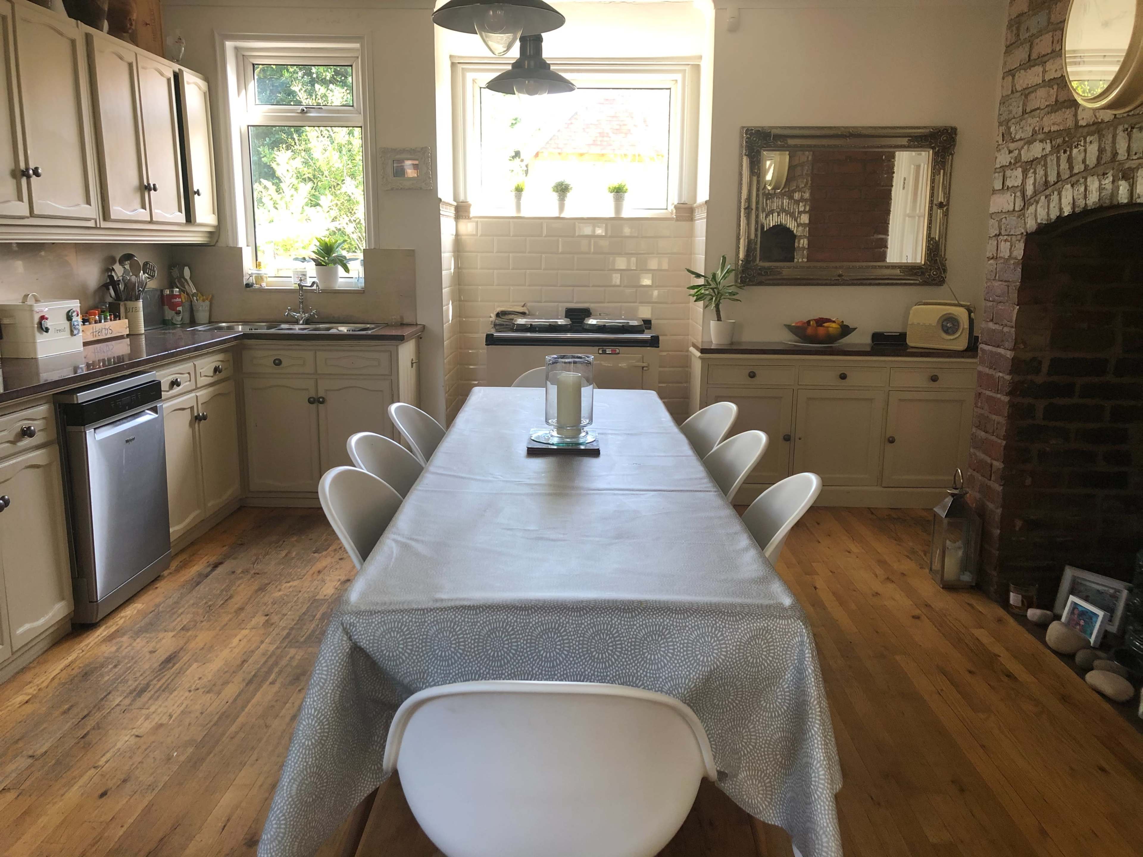 A spacious kitchen features a long dining table with white chairs, wooden floors, and cabinets along the walls, illuminated by natural light from the windows.