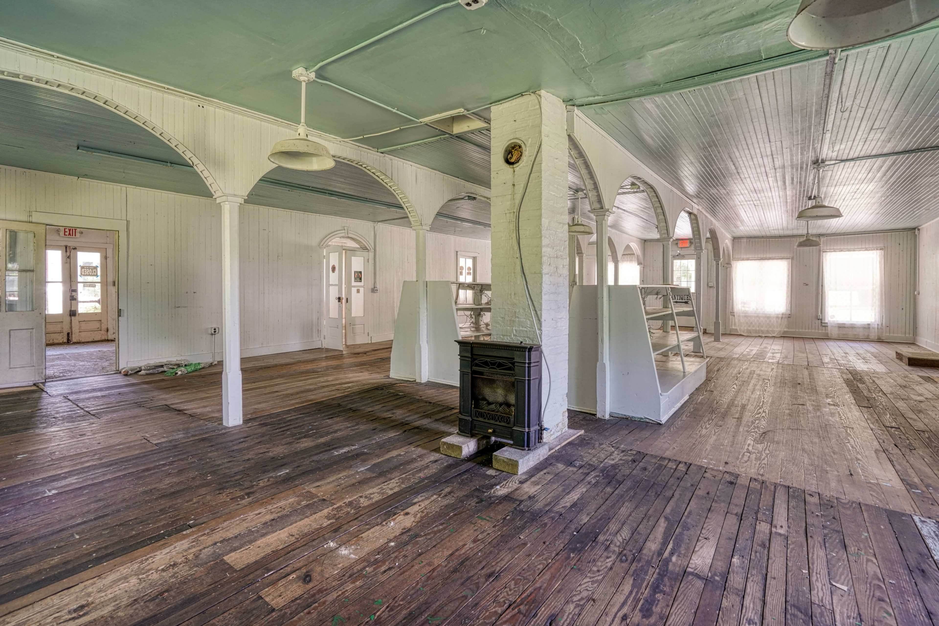 The image shows an empty interior space with wooden floors, white walls, and several arched pillars, featuring a decorative fireplace in the center.