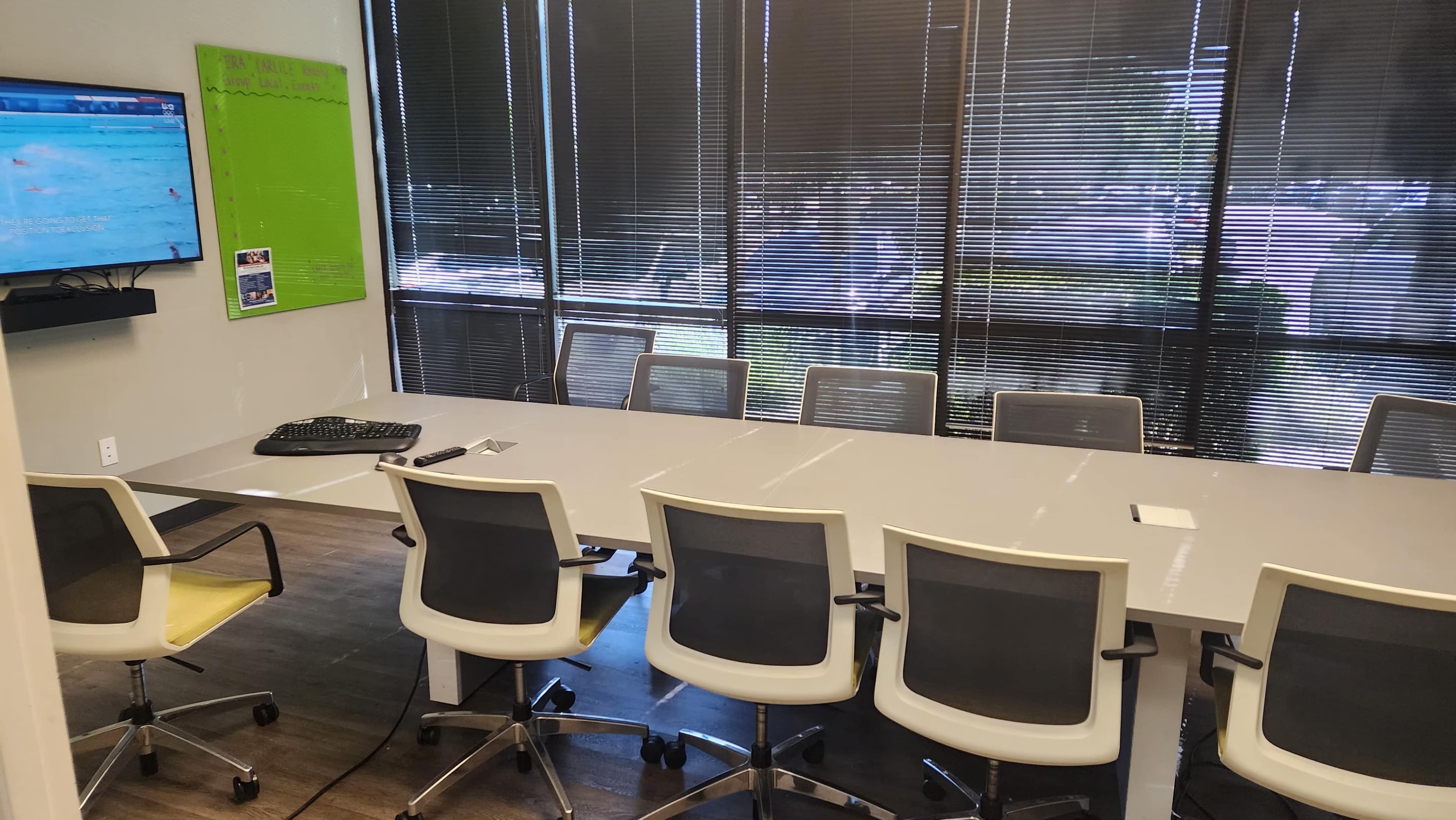 A modern conference room features a long table surrounded by ergonomic chairs, with a large screen displaying content and large windows lining one wall.
