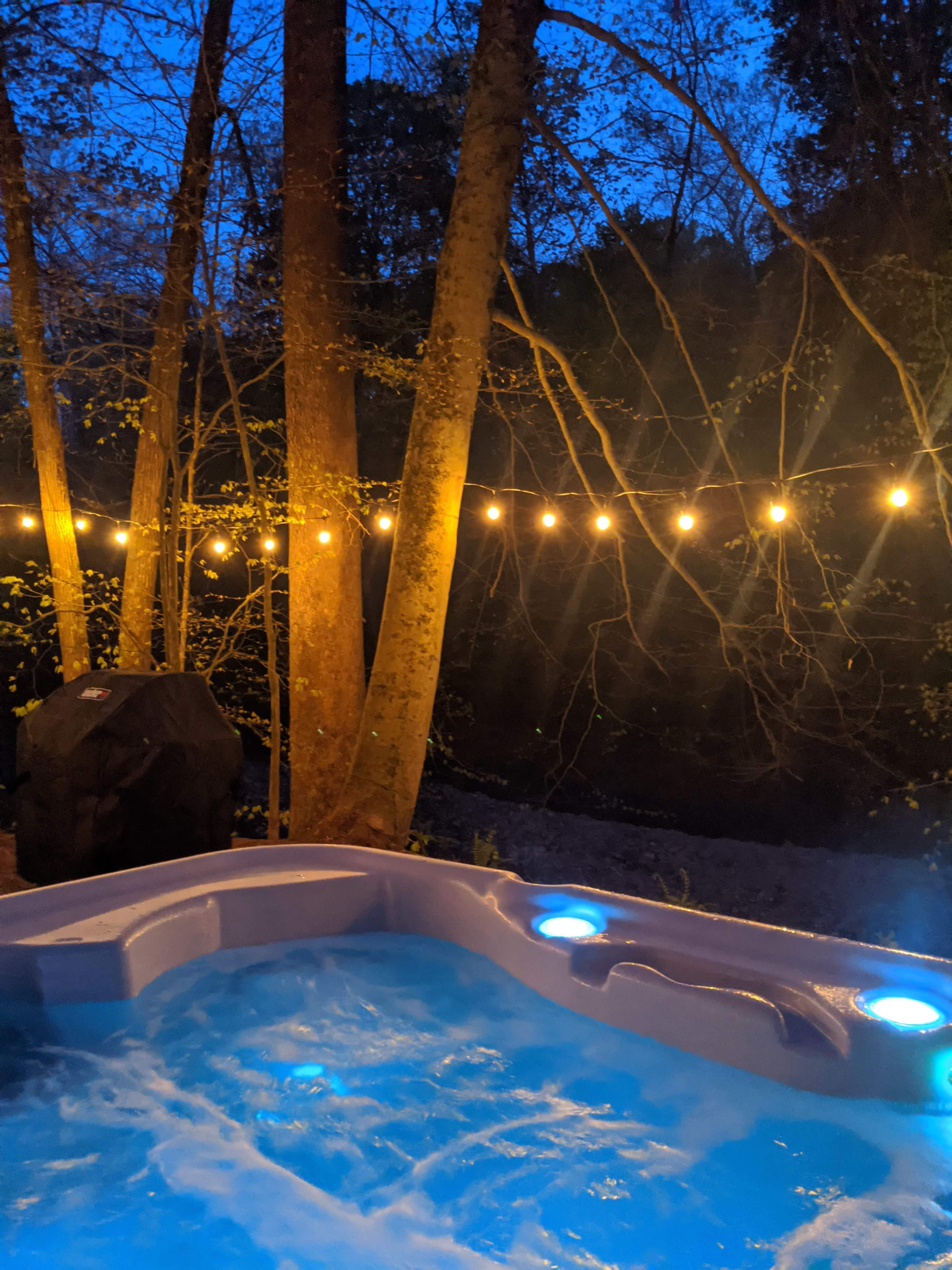 A hot tub illuminated by blue lights, surrounded by trees and a string of warm, glowing lights against a twilight sky.