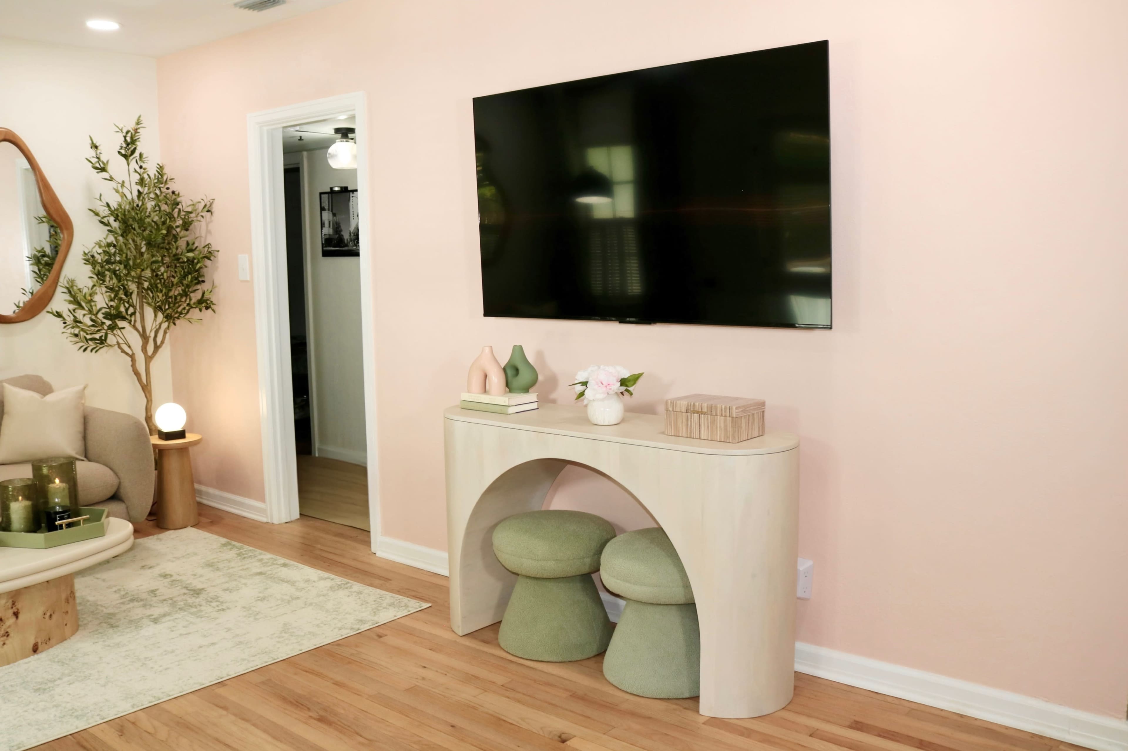 The room features a light pink wall with a large TV mounted above a pale wood console table, flanked by two green stools.