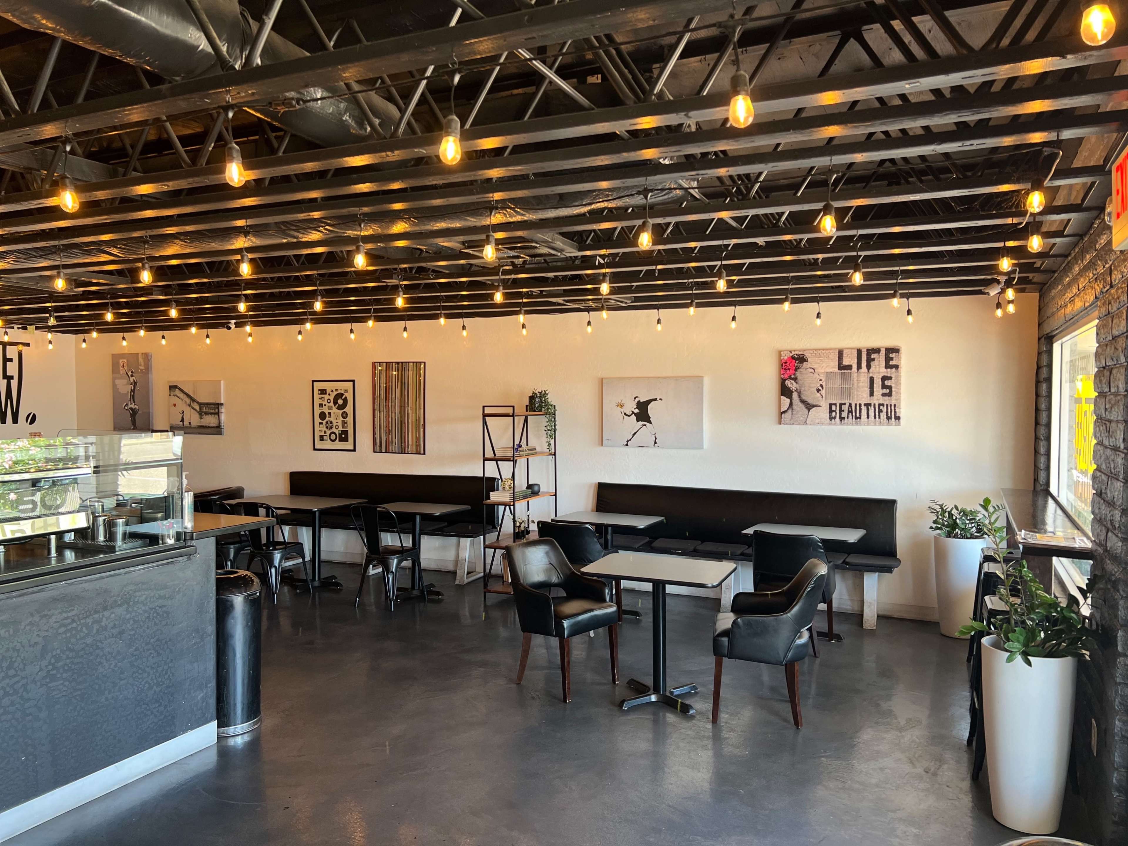 An interior of a café with a seating area featuring black benches, tables, and wall art, illuminated by hanging string lights.