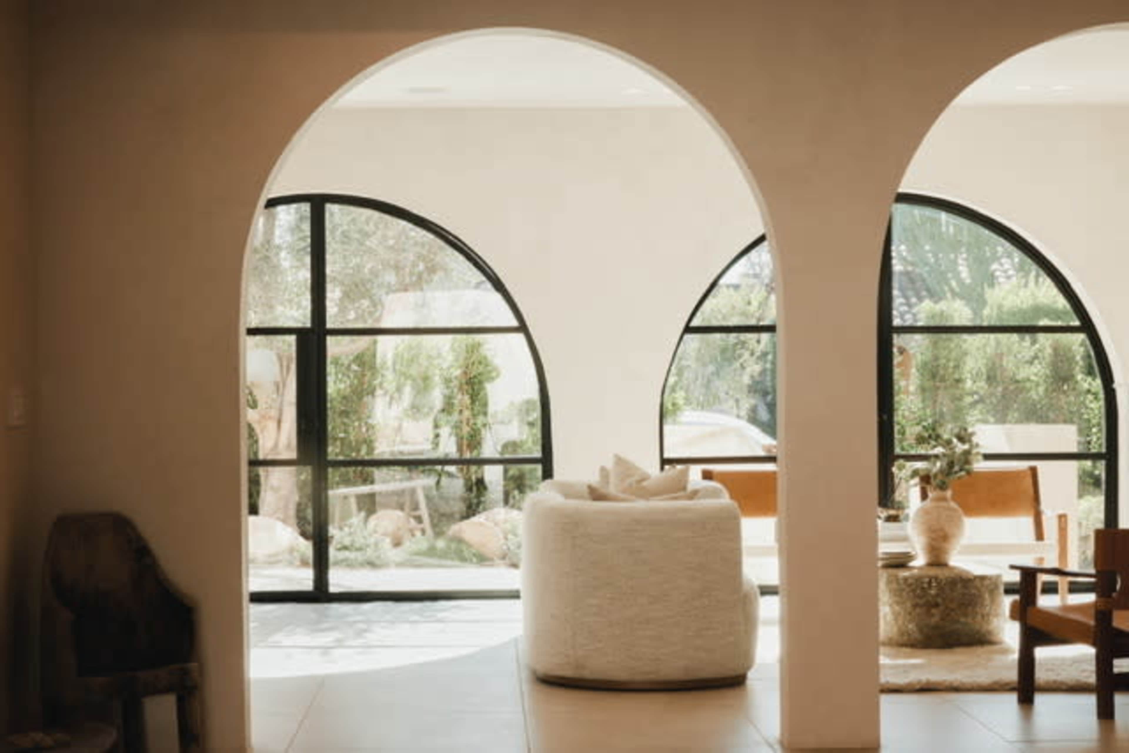 The image shows a living space featuring large arched windows, a round white upholstered chair, and wooden furniture, with plants visible outside.