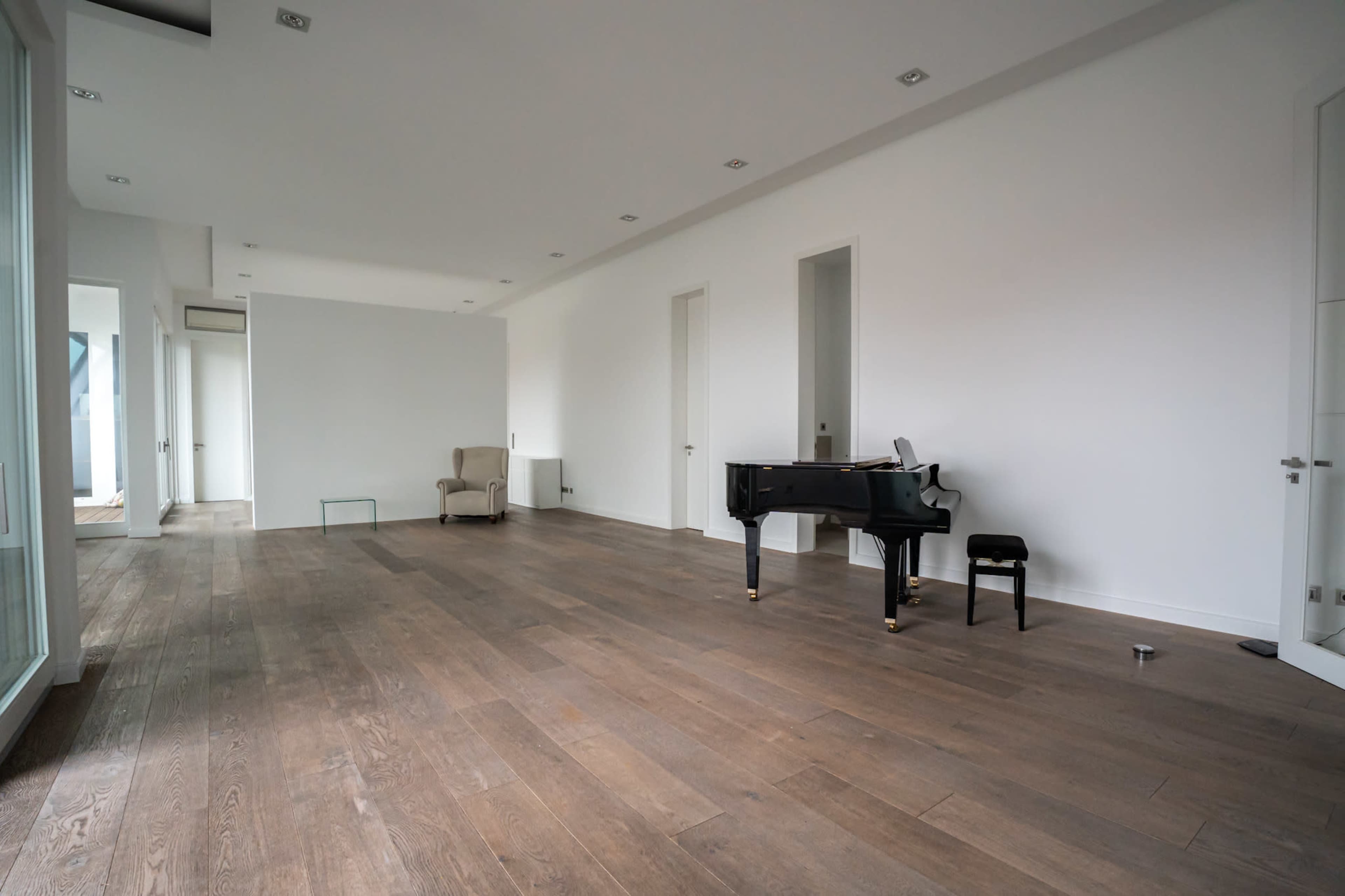 A spacious, empty room with a polished wooden floor, featuring a black grand piano and a beige armchair.