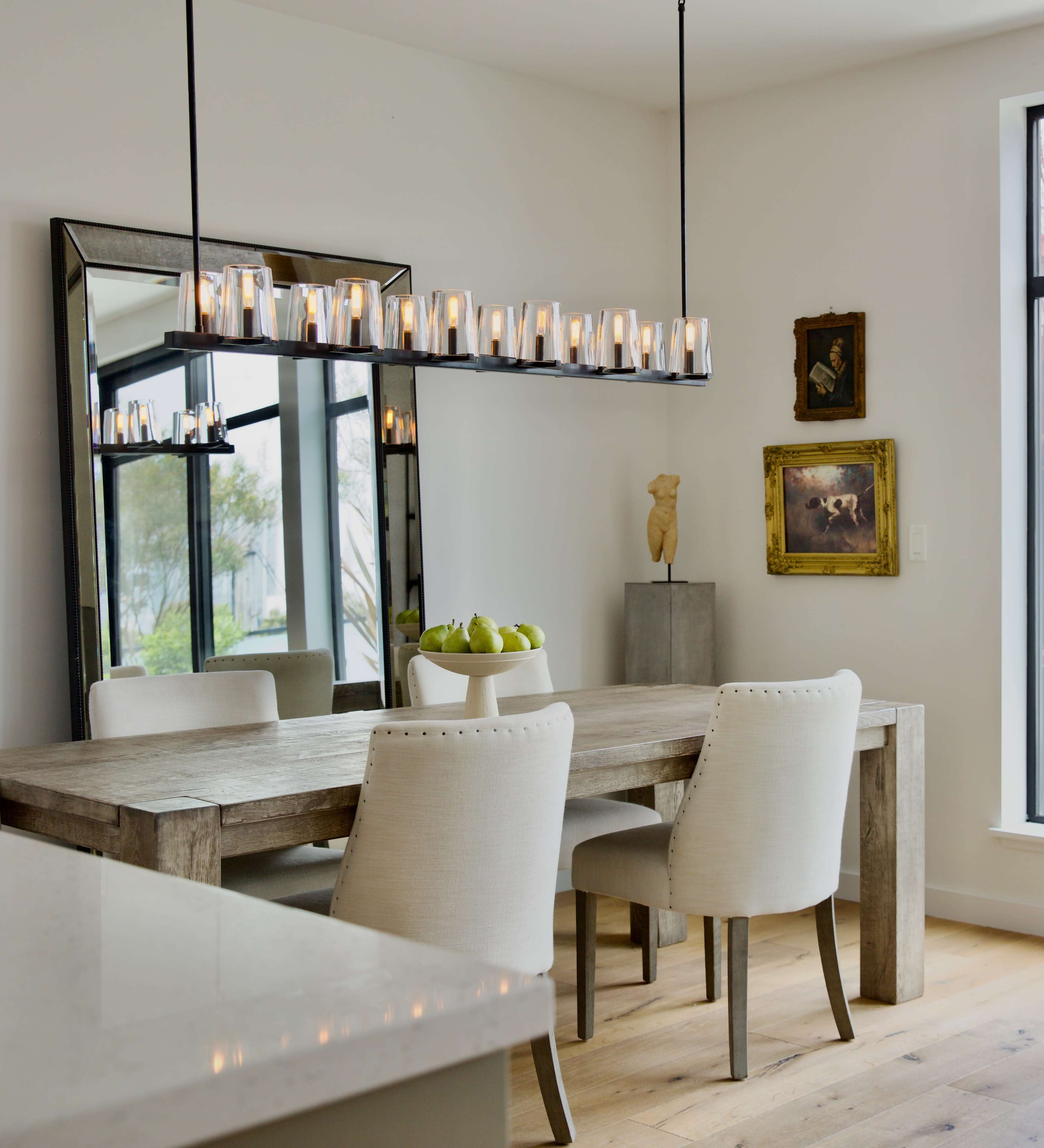 A modern dining area with a long wooden table, upholstered chairs, a contemporary chandelier, and a large mirror on the wall.