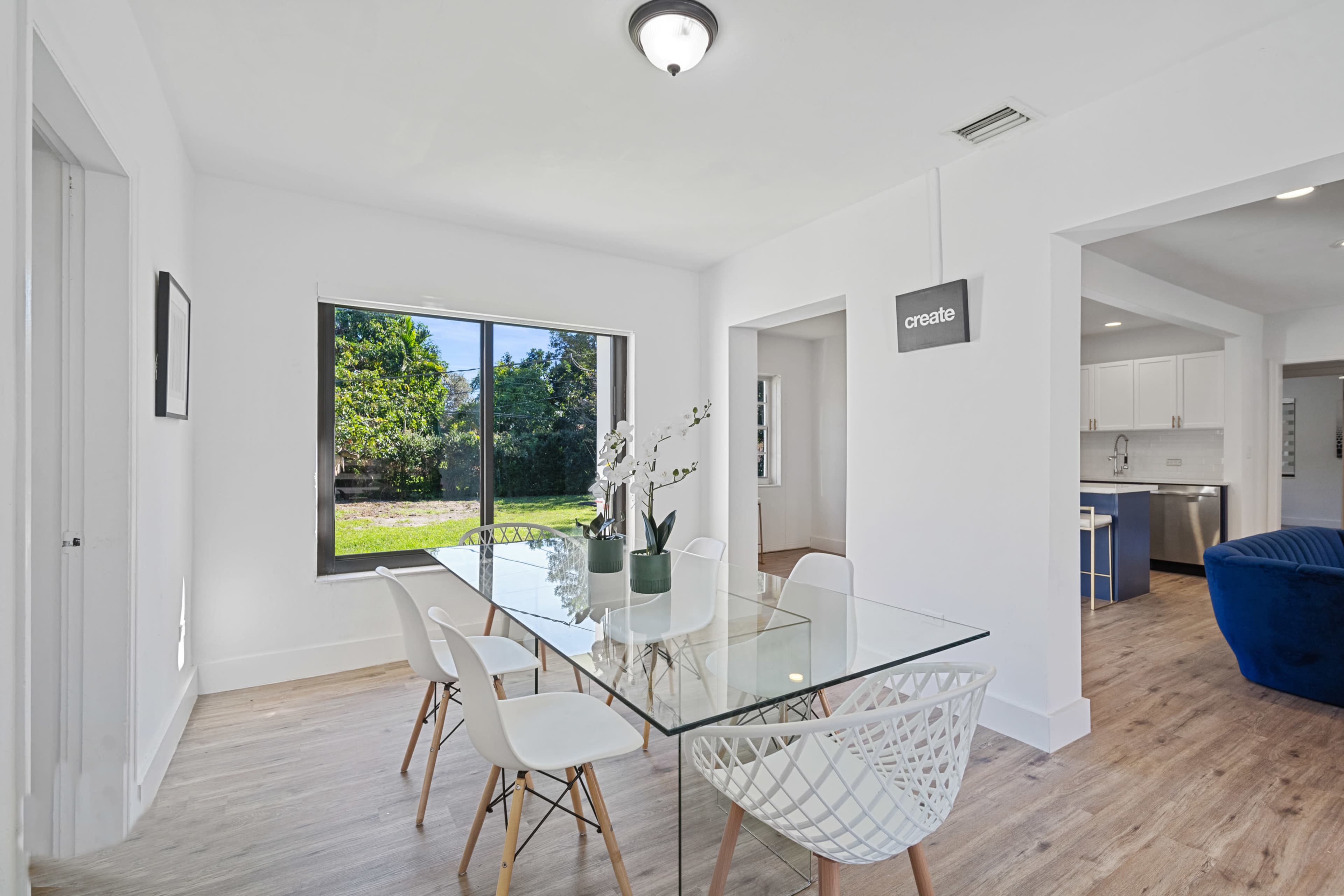 A modern dining area features a glass table surrounded by white chairs, with a view of a green outdoor space through a large window.