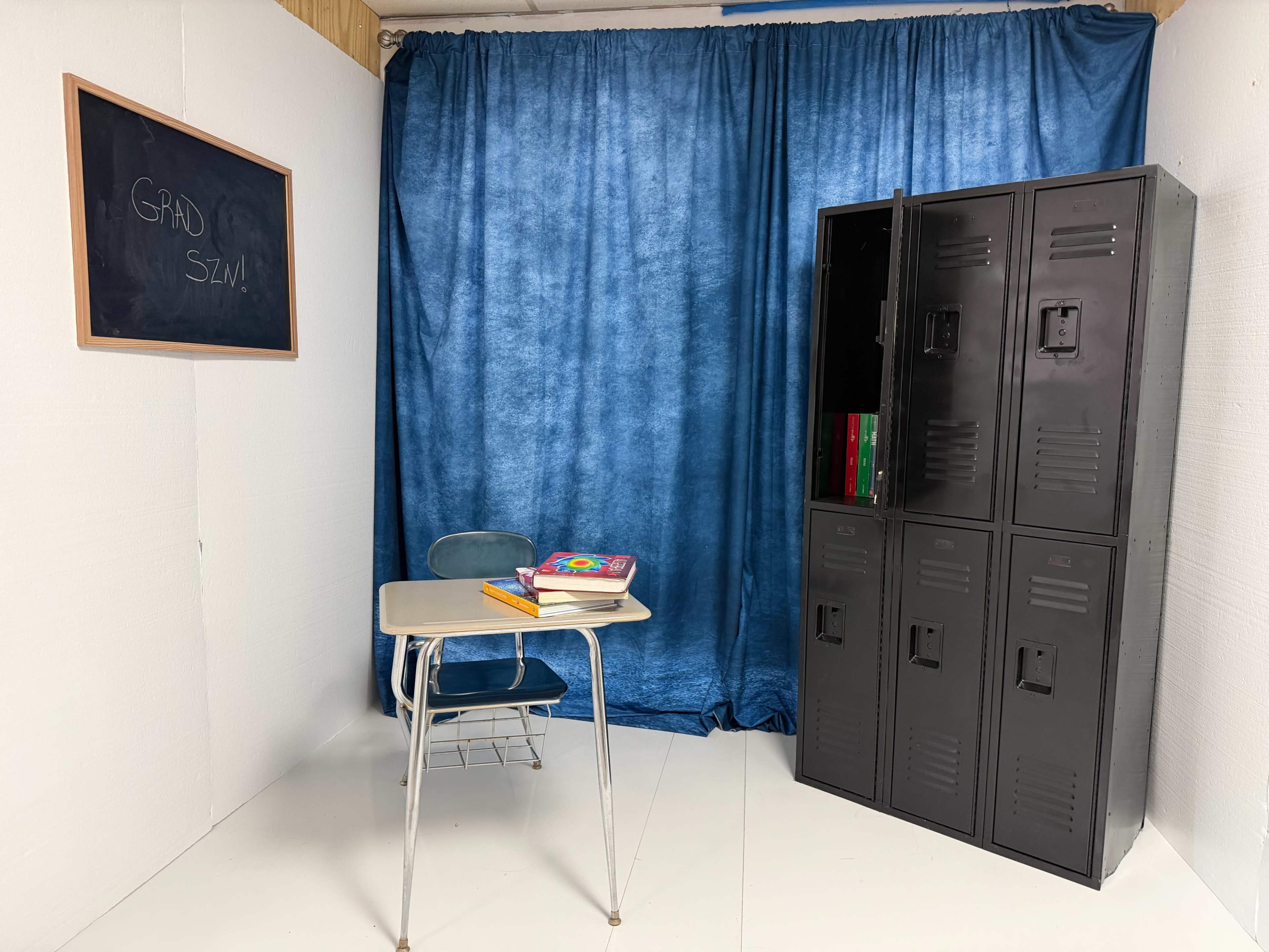 The image shows a small classroom-like setting with a blue curtain backdrop, a wooden desk with books, and a row of black lockers against the wall.