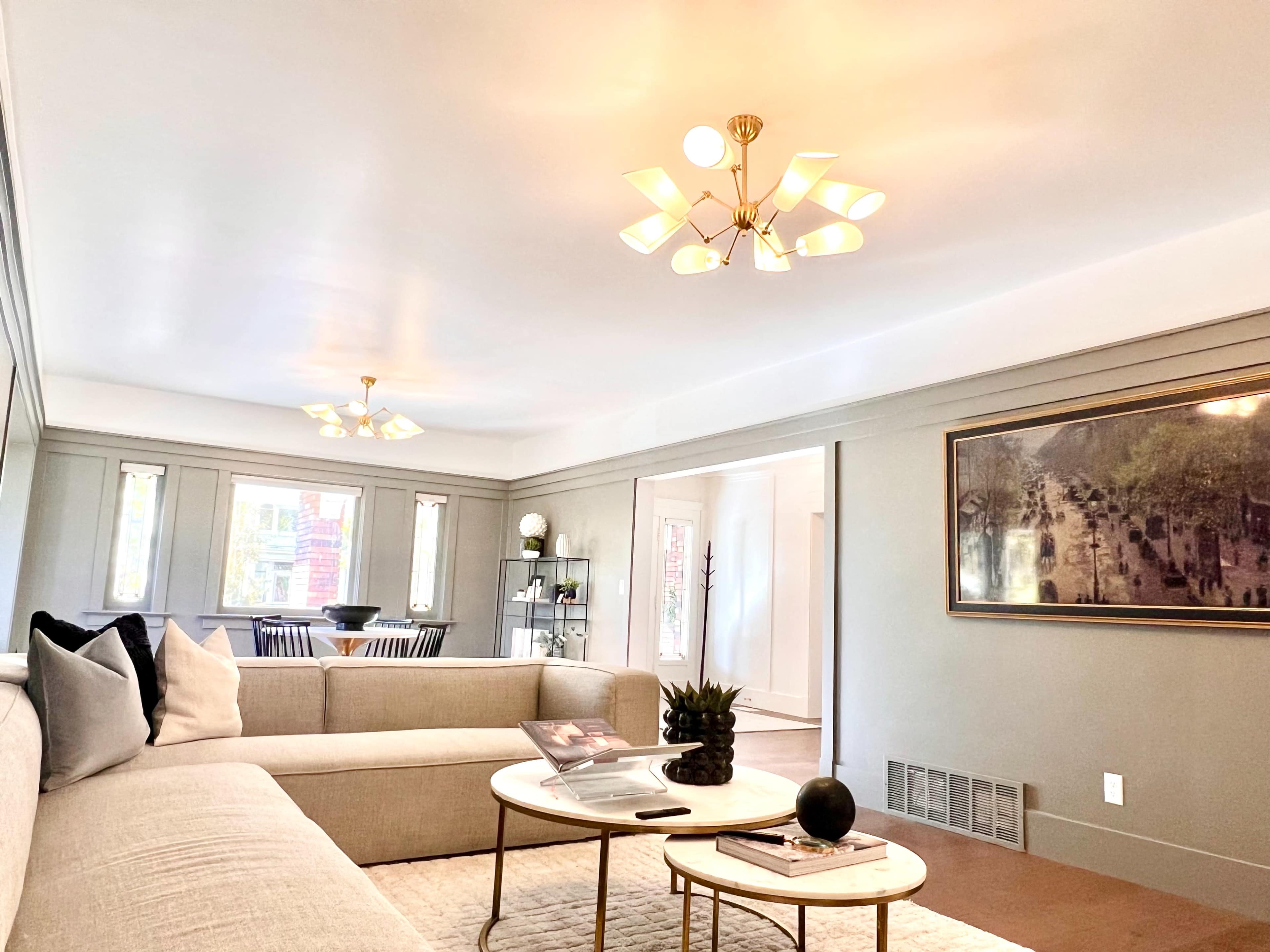 A modern living room features a light-colored sectional sofa, two coffee tables, and several windows illuminating the space, leading to a dining area with a visible table and chairs.