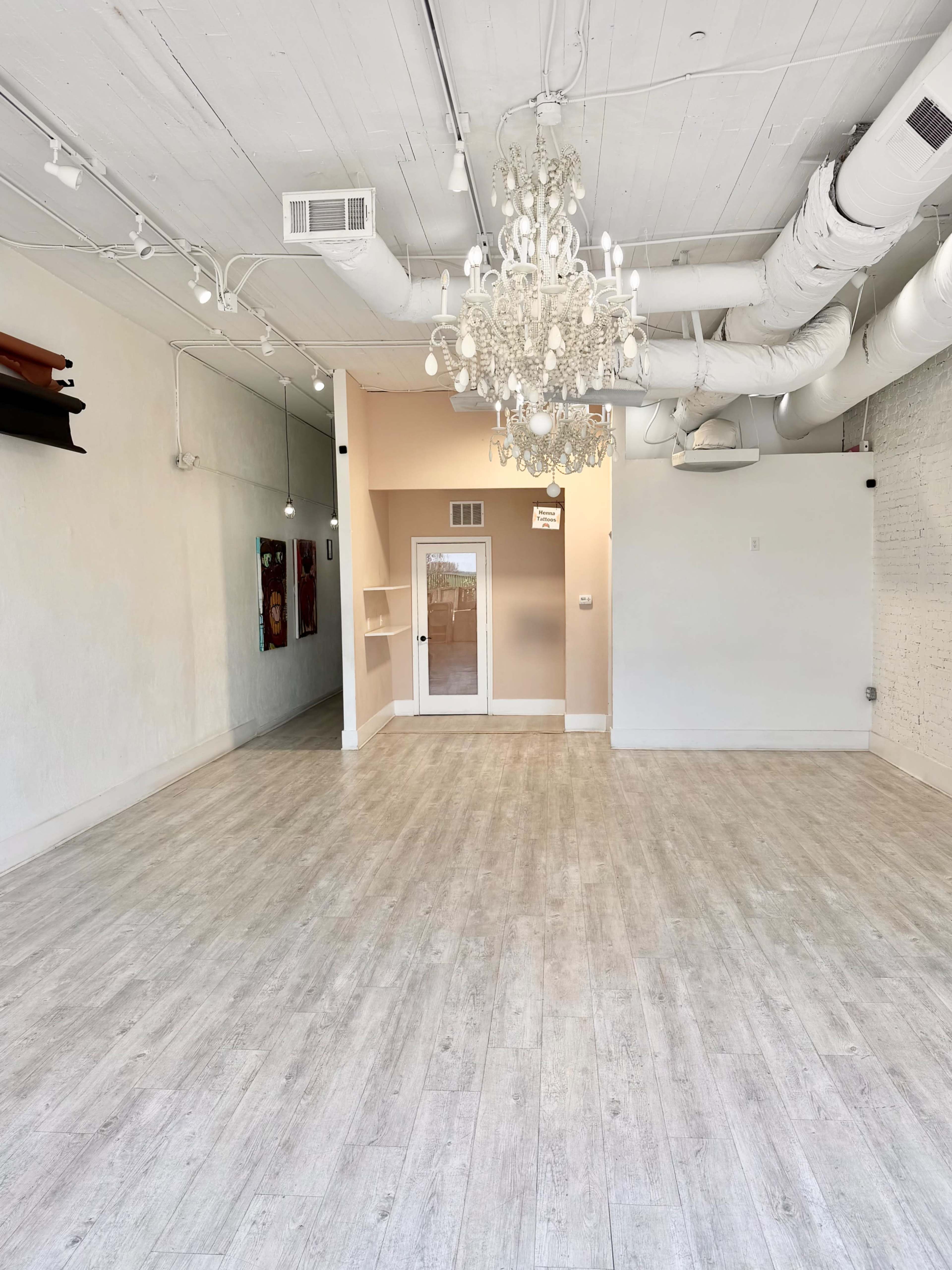 The image shows an empty interior space featuring light-colored wooden flooring, white walls, and a chandelier hanging from the ceiling.