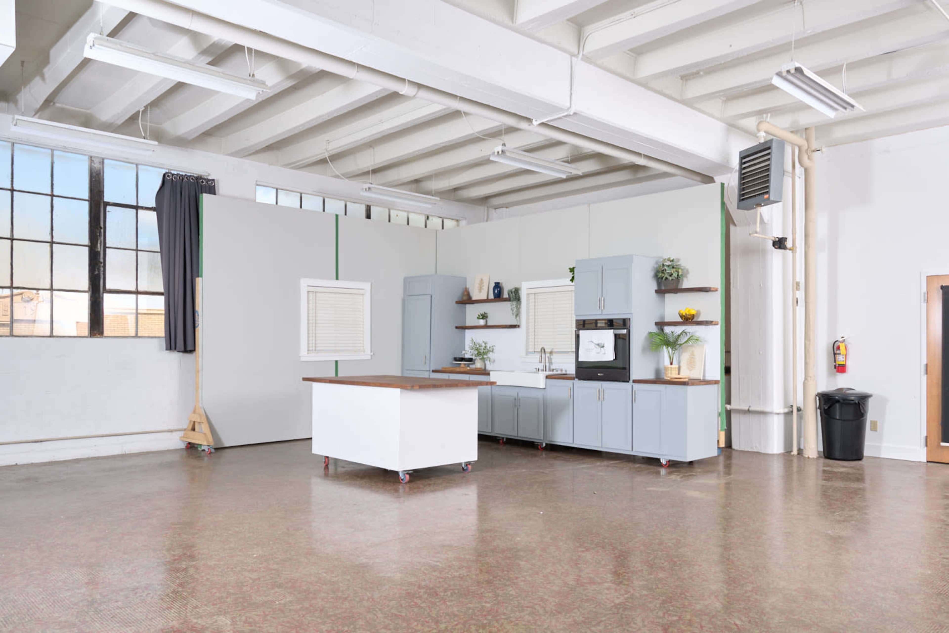 The image shows an open, spacious kitchen area with blue cabinetry, a central island, and large windows allowing natural light to enter.