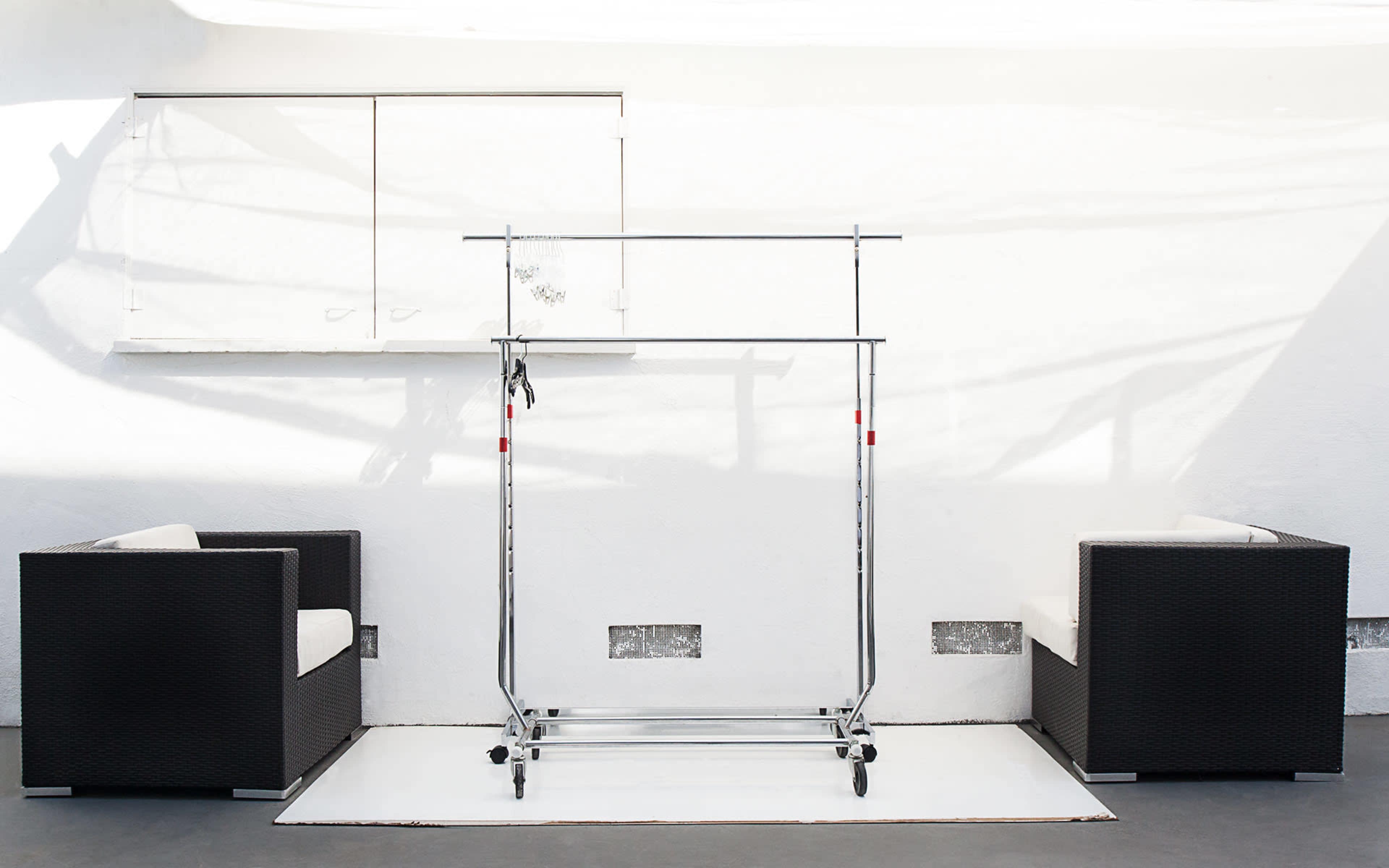 A minimalist space with two woven chairs flanking a clothing rack against a white wall.