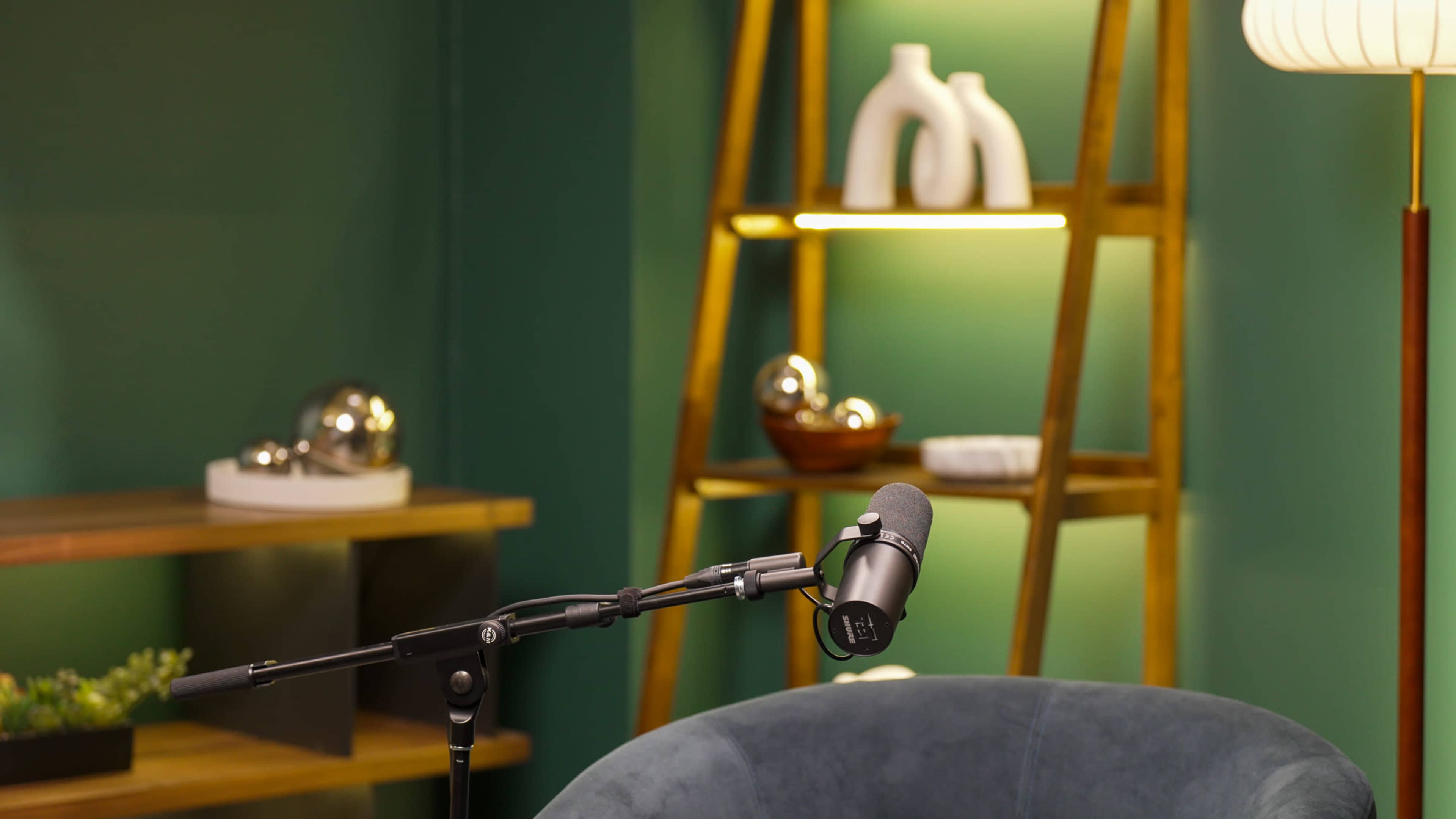 A microphone is positioned on a stand next to a modern chair, with a backdrop of shelves displaying various decorative items.
