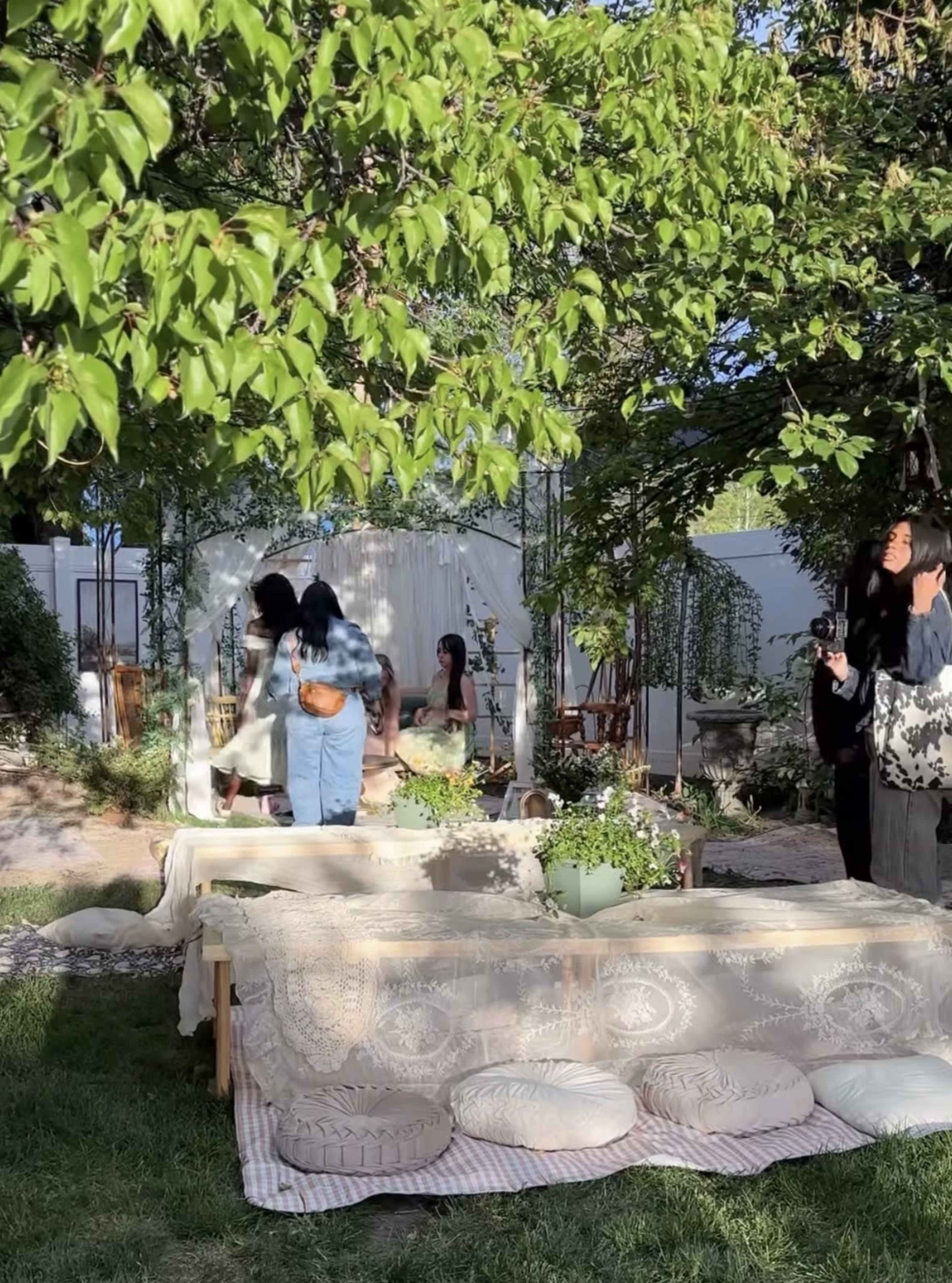 A group of people gather in a garden setting adorned with wooden furniture and greenery, while one person takes photographs.