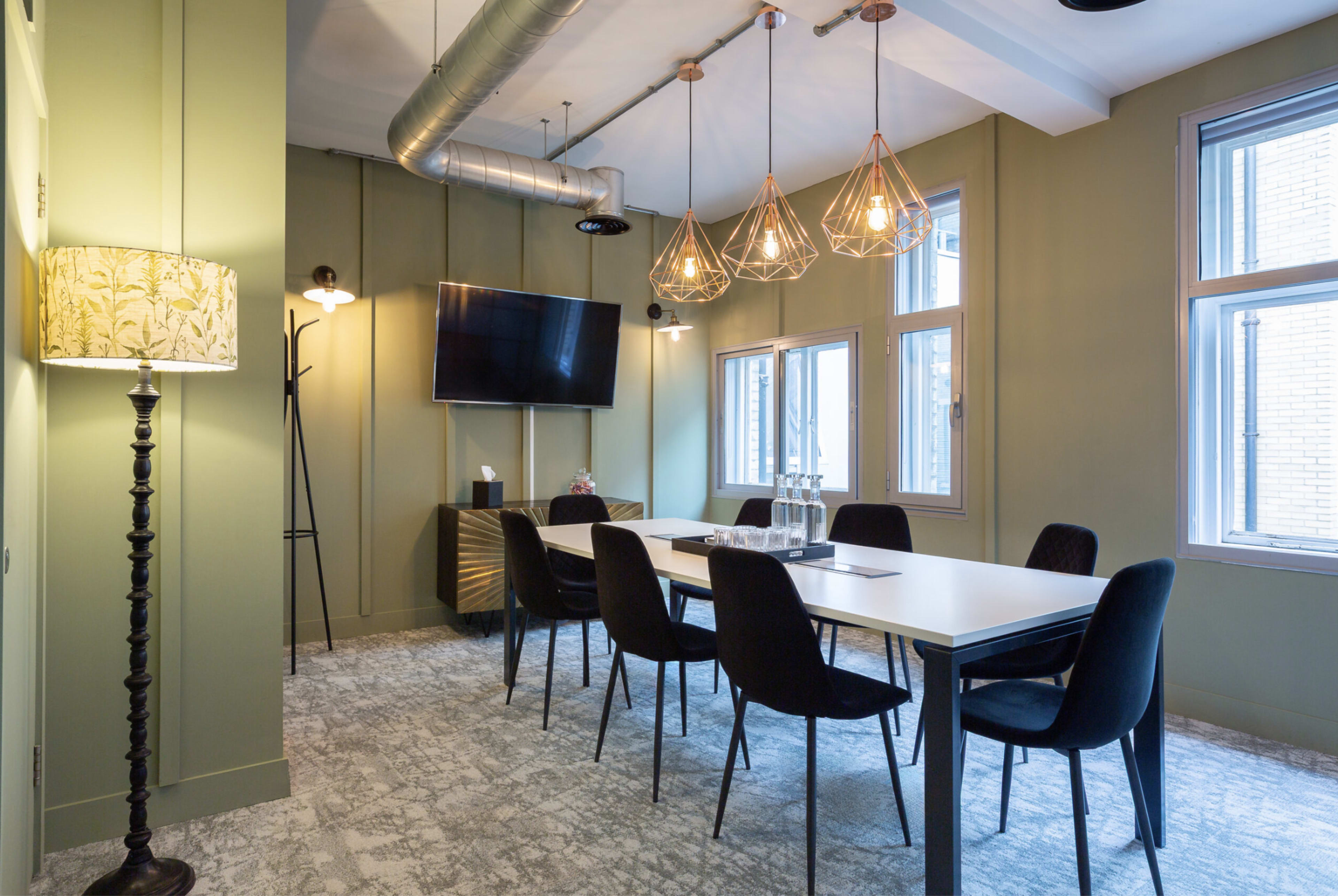 A modern conference room features a long white table surrounded by black chairs, with large windows and stylish lighting fixtures overhead.