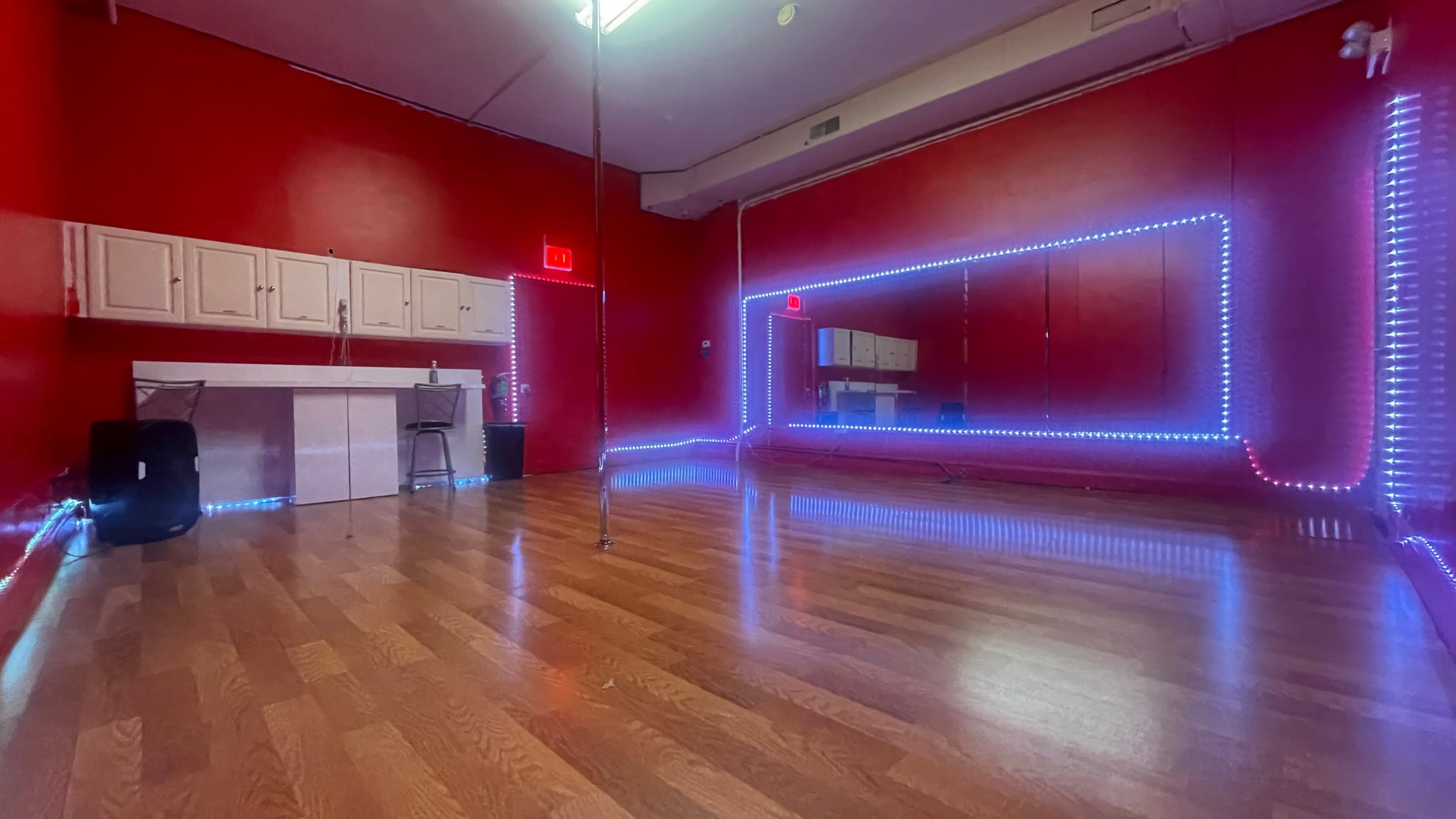 A dance studio features a polished wooden floor with red walls, illuminated by blue LED lights along the mirrors and perimeter.