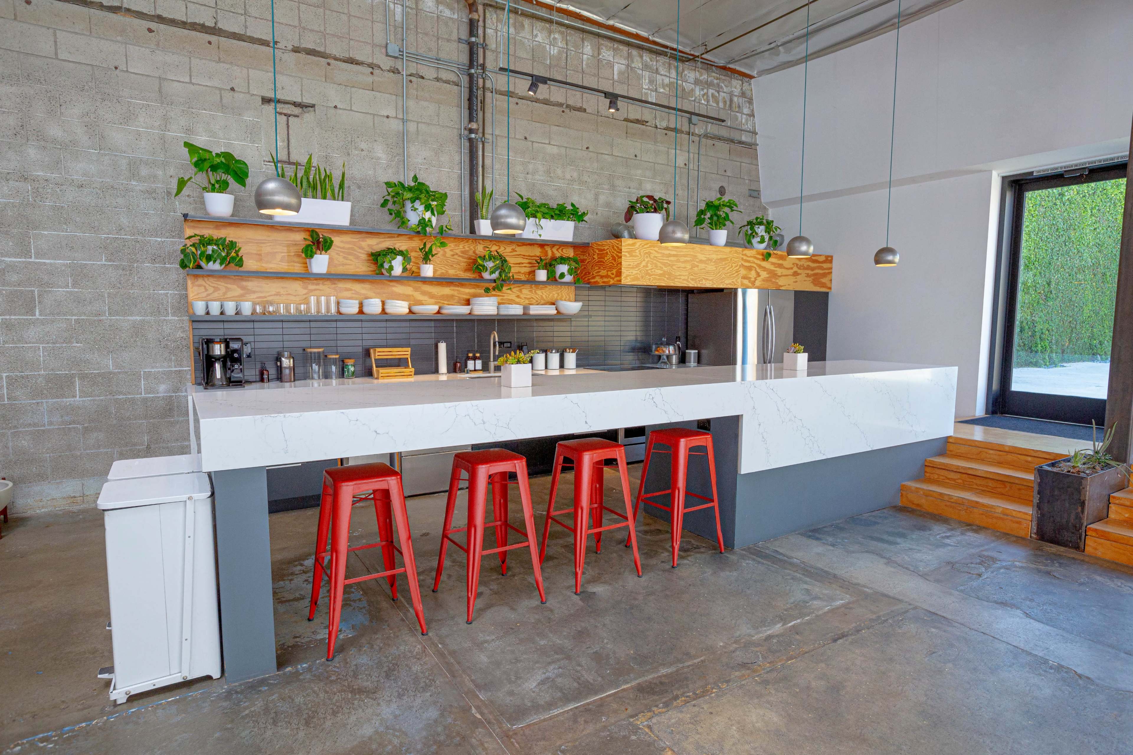 A modern kitchen bar with a white marble countertop, wooden shelving adorned with plants, and red metal stools.