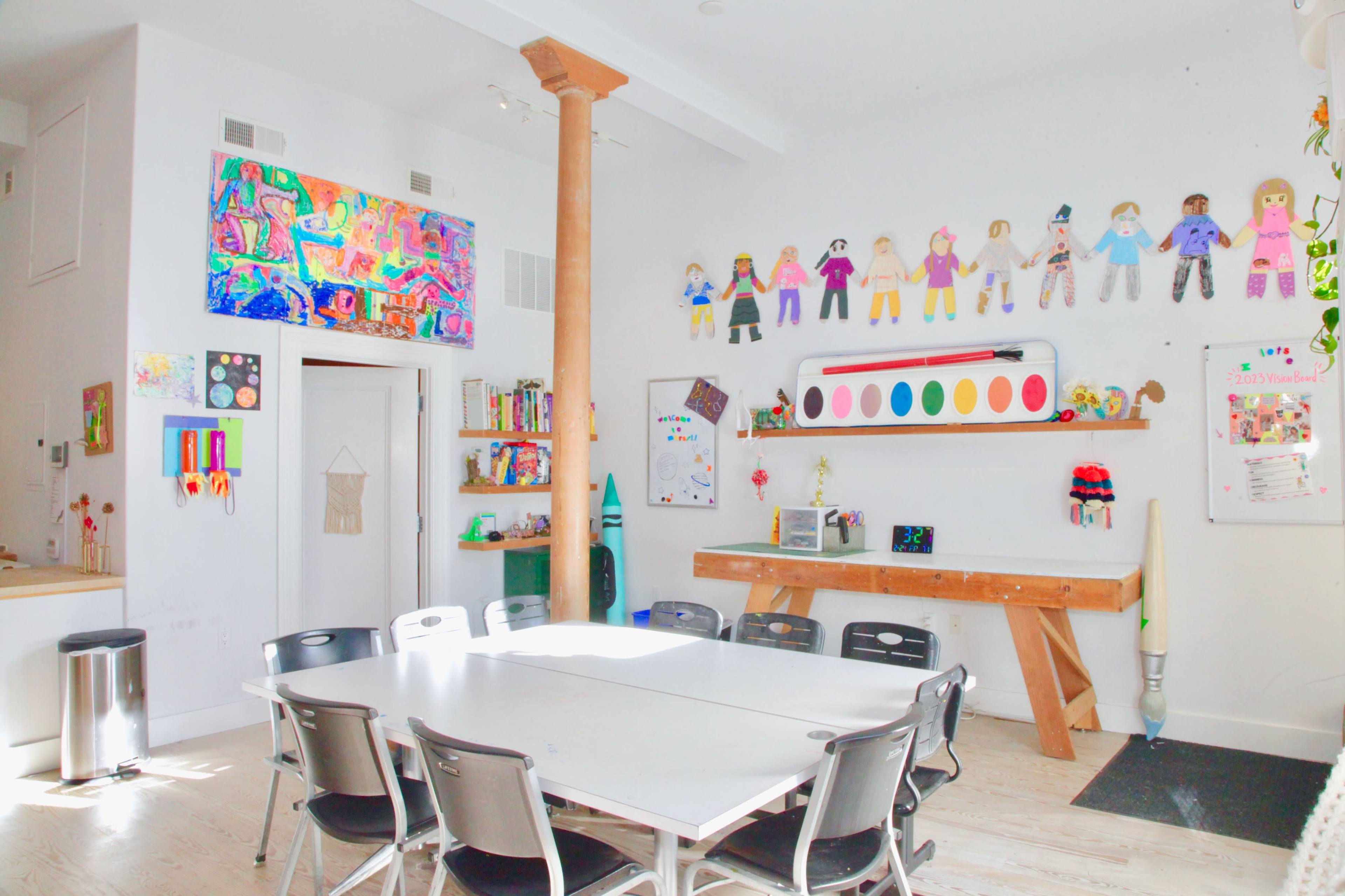 A brightly lit classroom with a large table surrounded by chairs, colorful student artworks on the walls, and various teaching materials on shelves.
