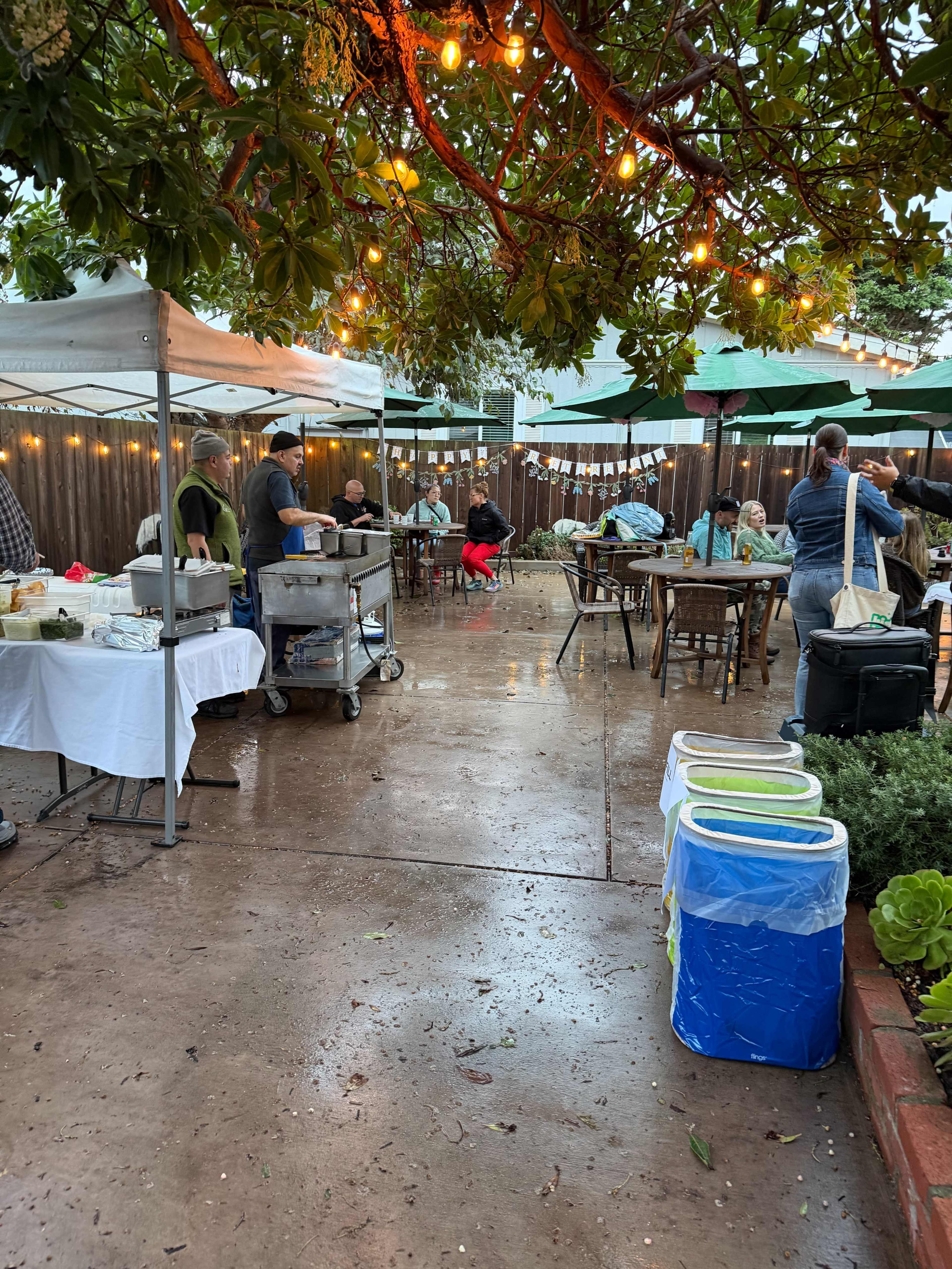 The image shows a patio area with tables set up under canopies, string lights overhead, and people gathered, some preparing food and others dining.