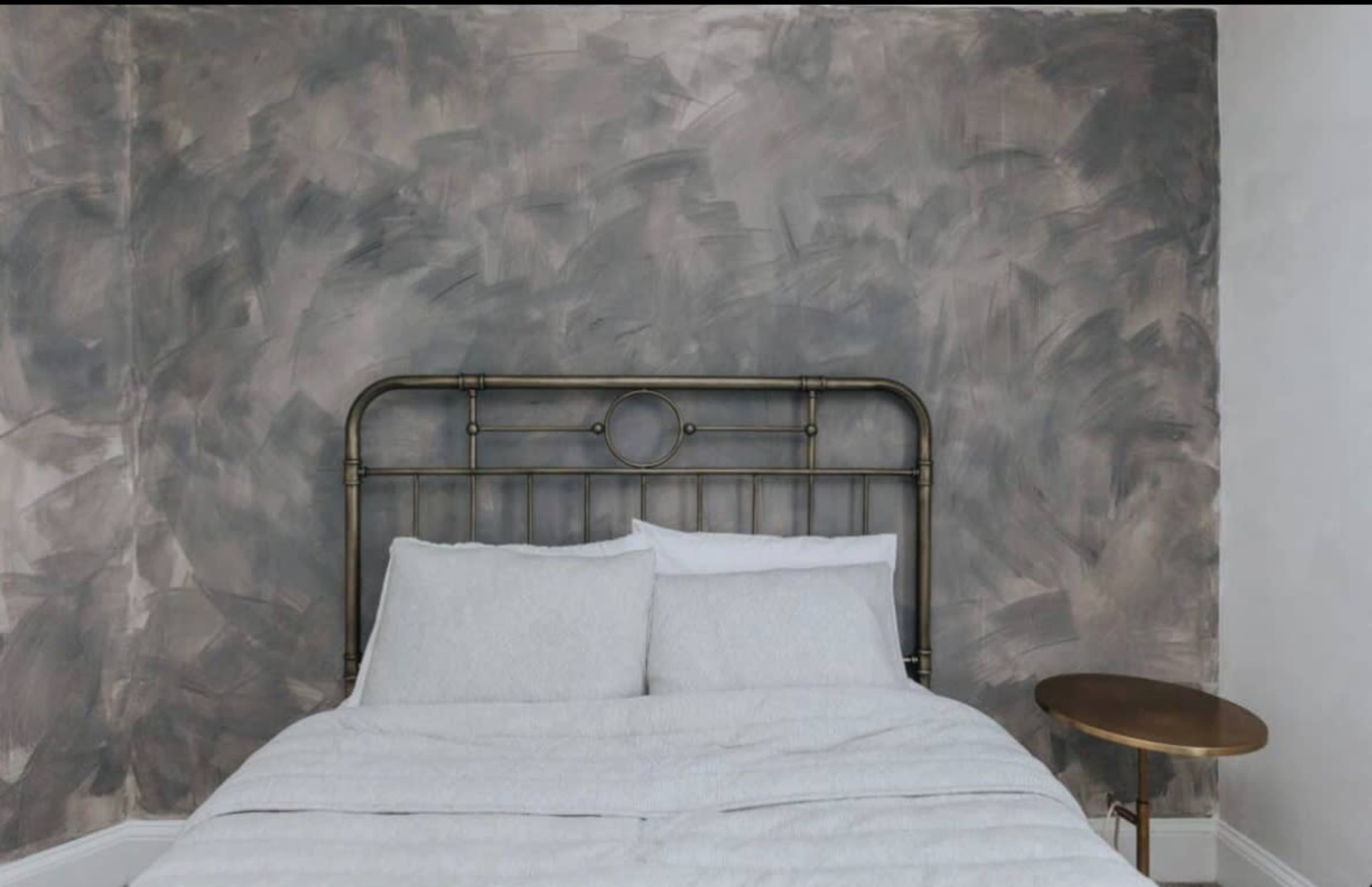 The image shows a bed with a metal headboard against a textured gray wall, accompanied by a small round side table.